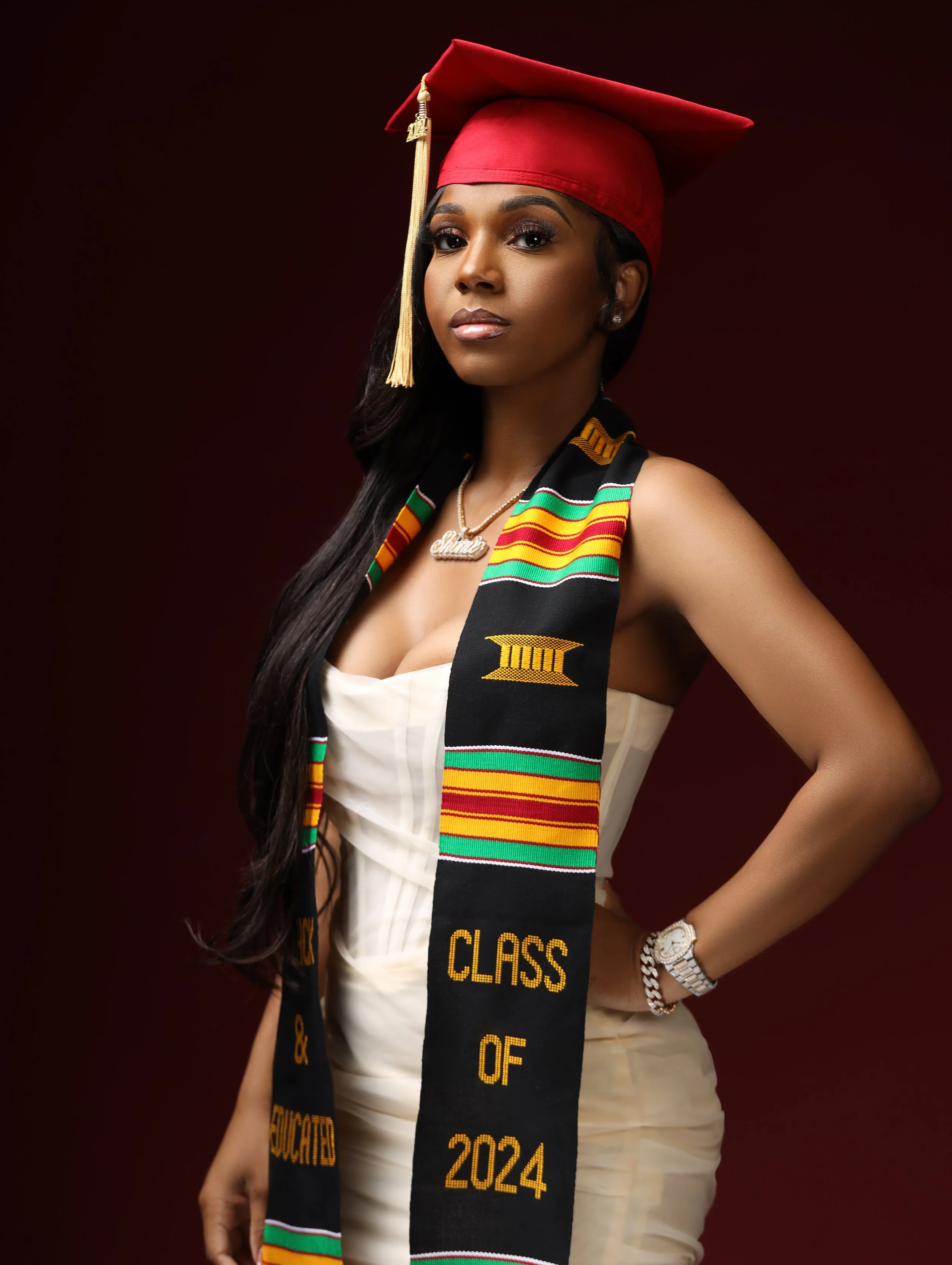 A young woman in a white strapless dress wearing a red graduation cap and mortarboard, with a colorful Kente cloth stole that reads 'Class of 2024' and 'Graduated', posing against a dark red background.