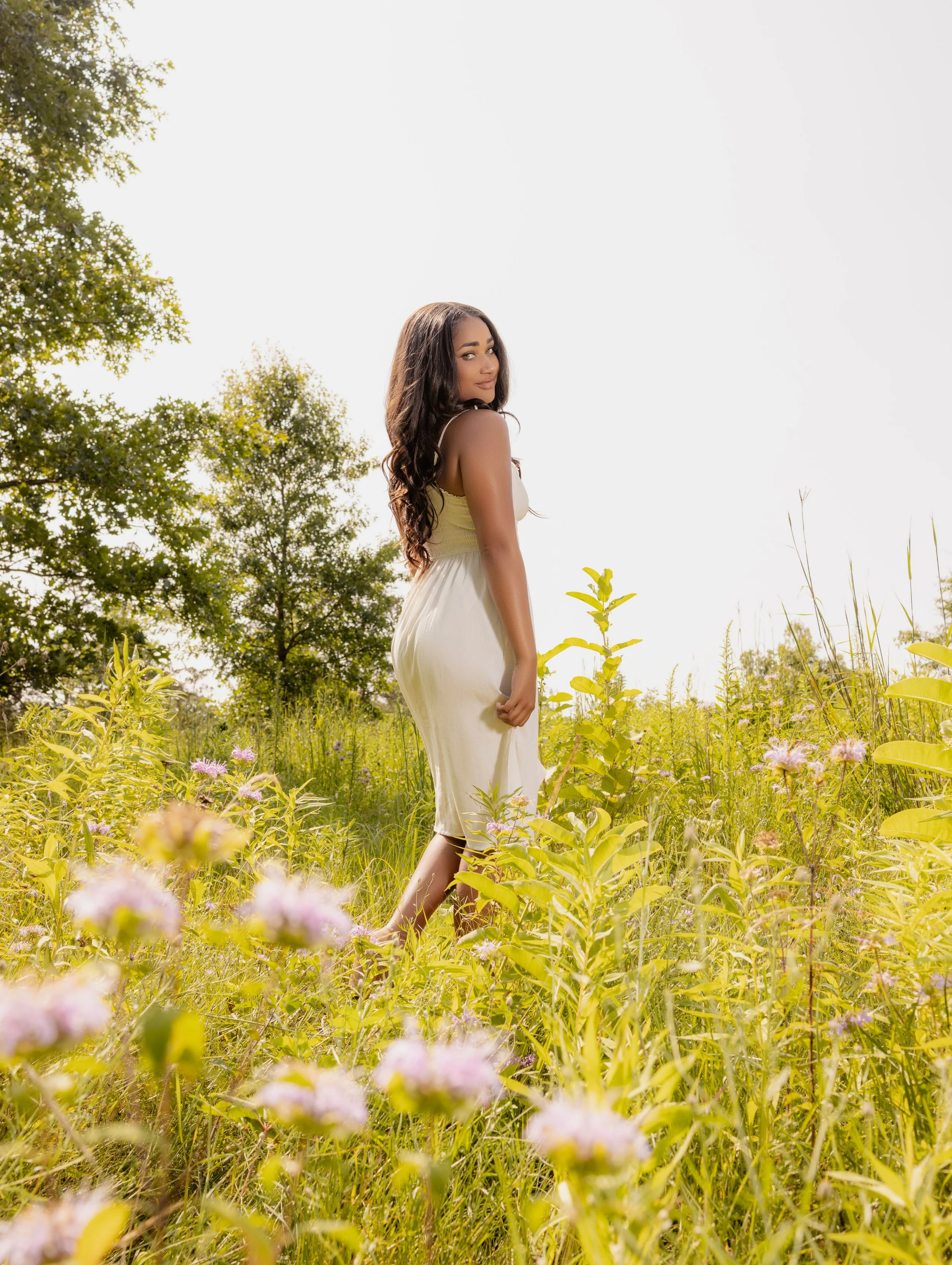 A woman with long, dark hair wearing a light yellow dress standing in a green meadow with flowering plants and trees in the background.