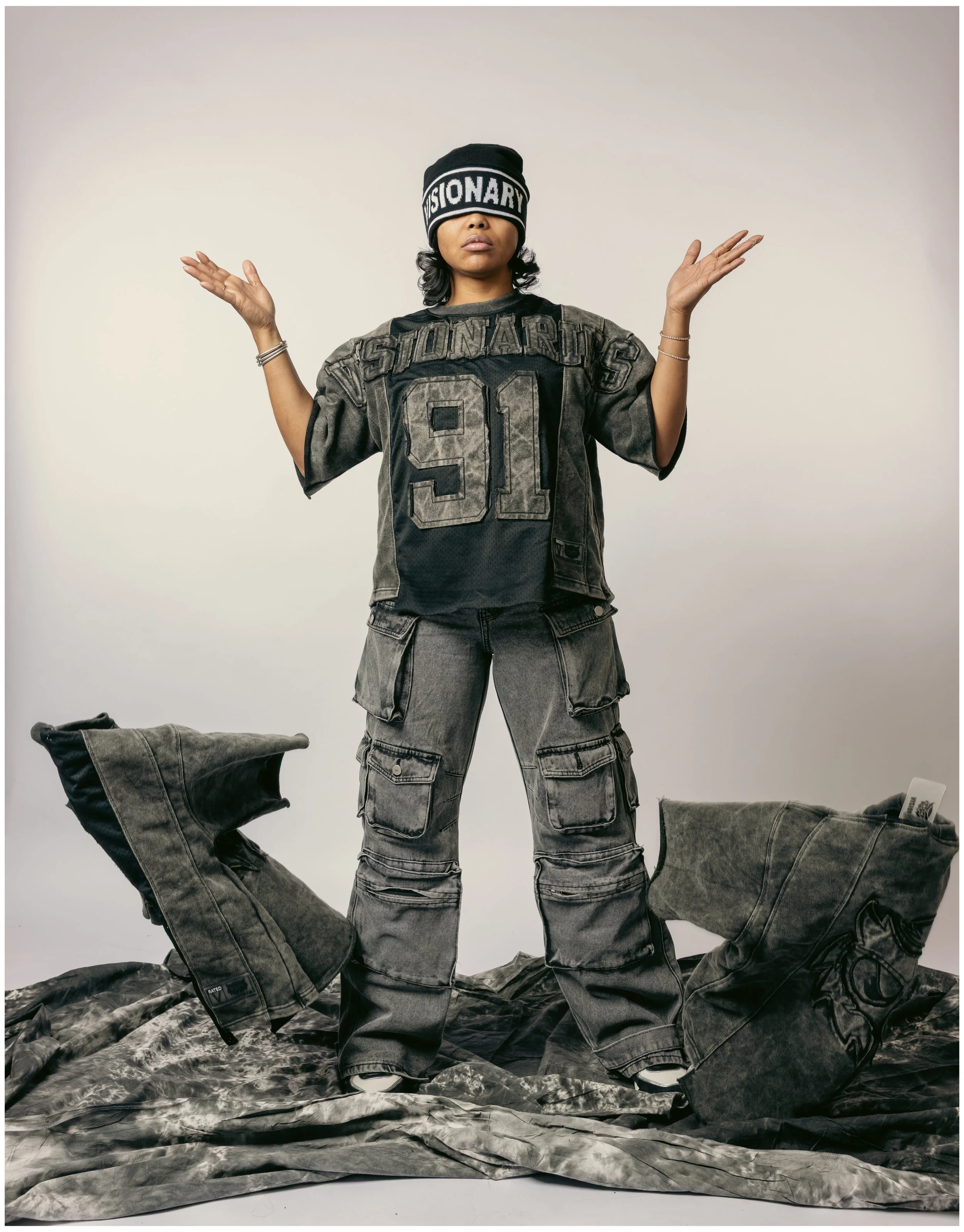 Woman wearing a black beanie with white lettering, a black and gray oversized sports jersey with the number 91 and the word 'STO NARICS,' gray cargo pants, standing on a fabric surface with supplement bags on either side.