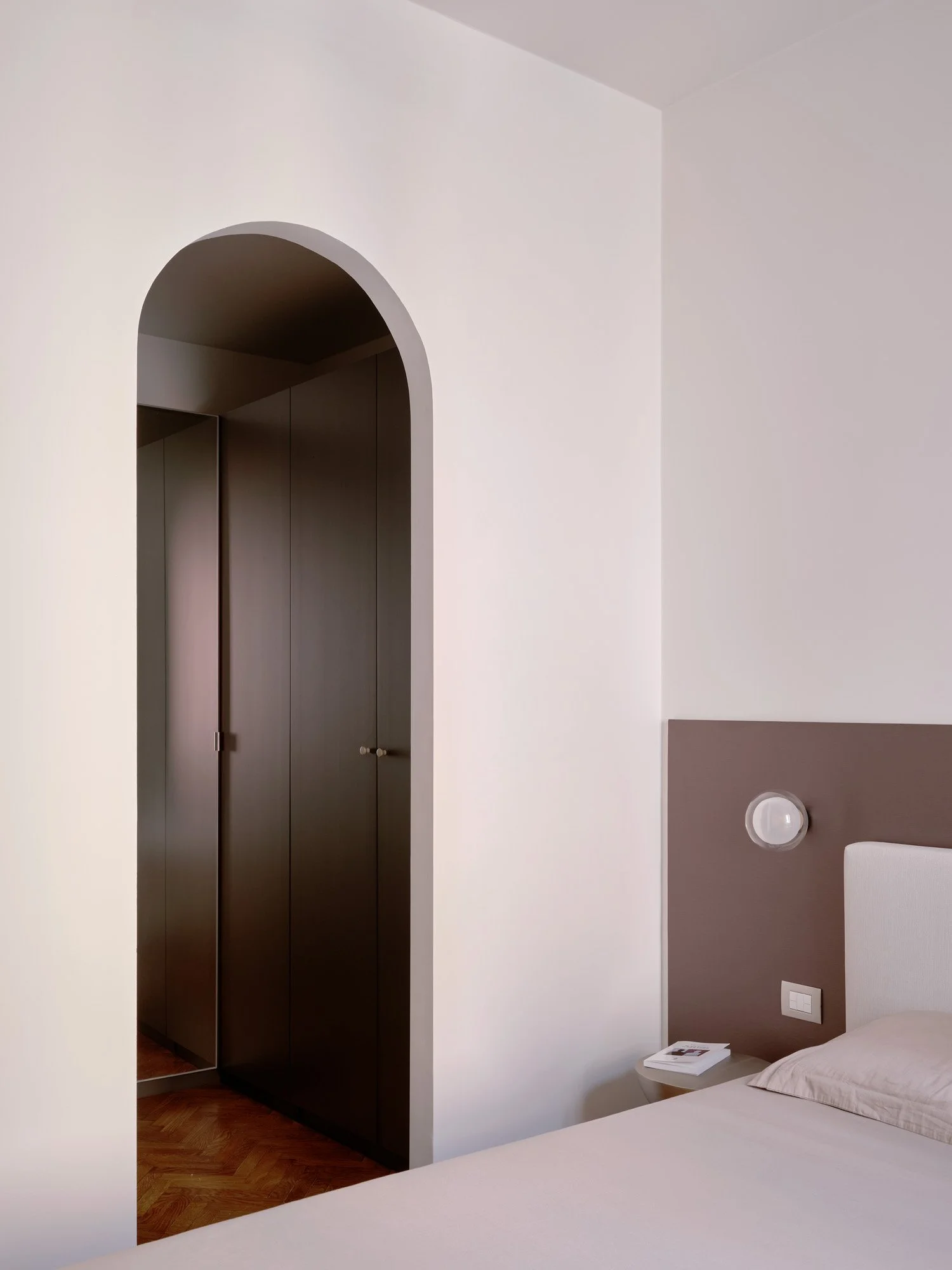 Camera da letto con pareti bianche, letto con biancheria beige, lampada da parete, comodino con libro, parete con pannello marrone e porta accanto a un armadio scuro con porta a battente.