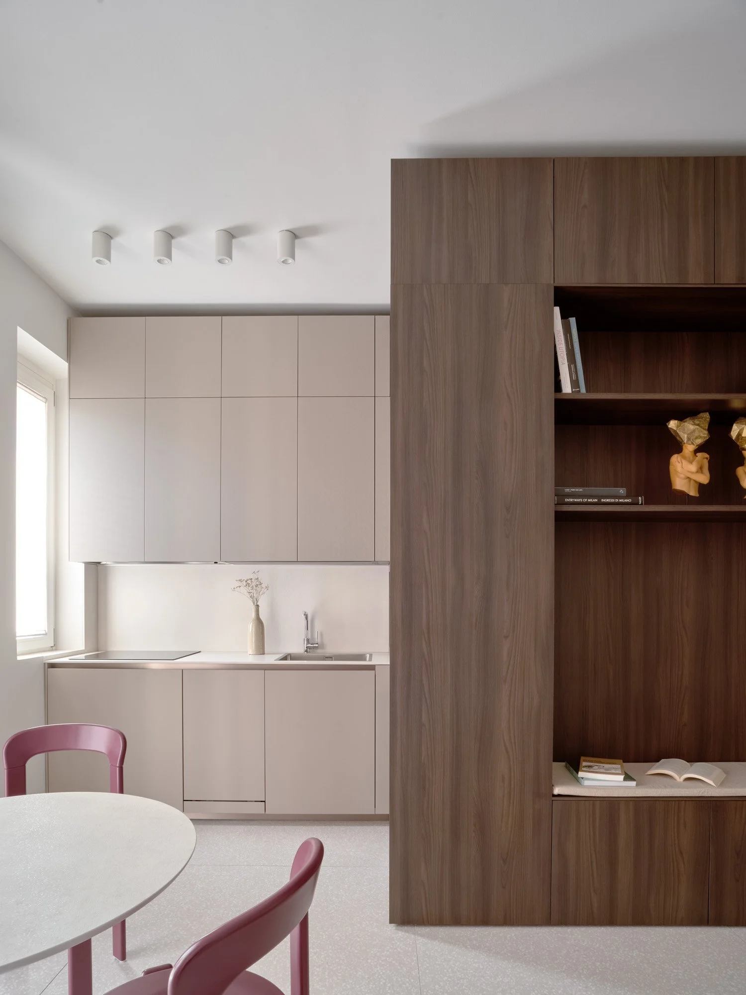 Cucina minimalista con mobili beige e rivestimento in legno, tavolo rotondo con sedie rosa, scaffale in legno con libri e decorazioni, finestra luminosa.
