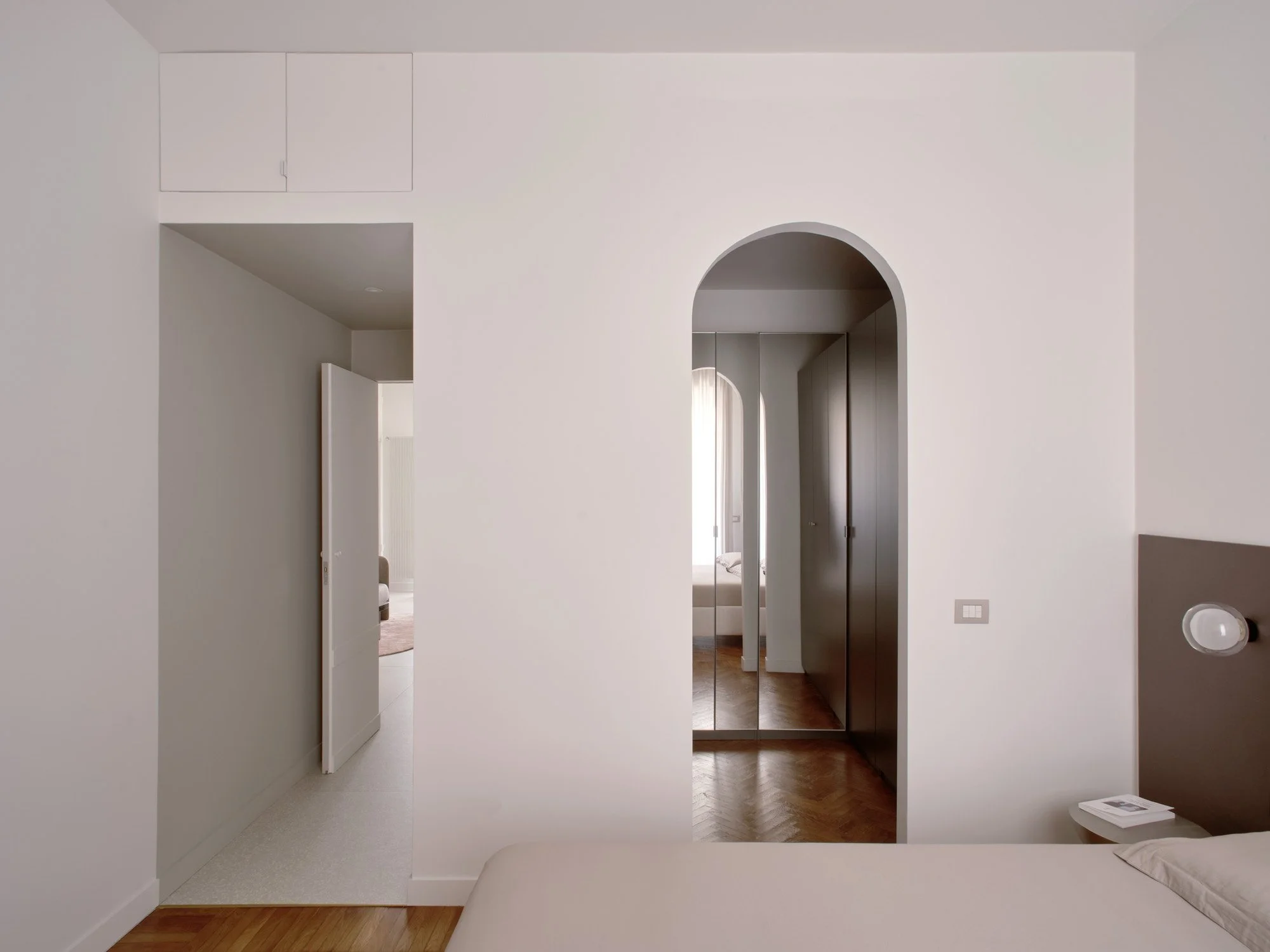 Camera con pareti bianche, letto, lampada da lettura e una piccola scrivania con un libro, ingresso a una stanza con armadio a specchio e porta aperta, vista sulla stanza successiva con letto e finestra con tende.