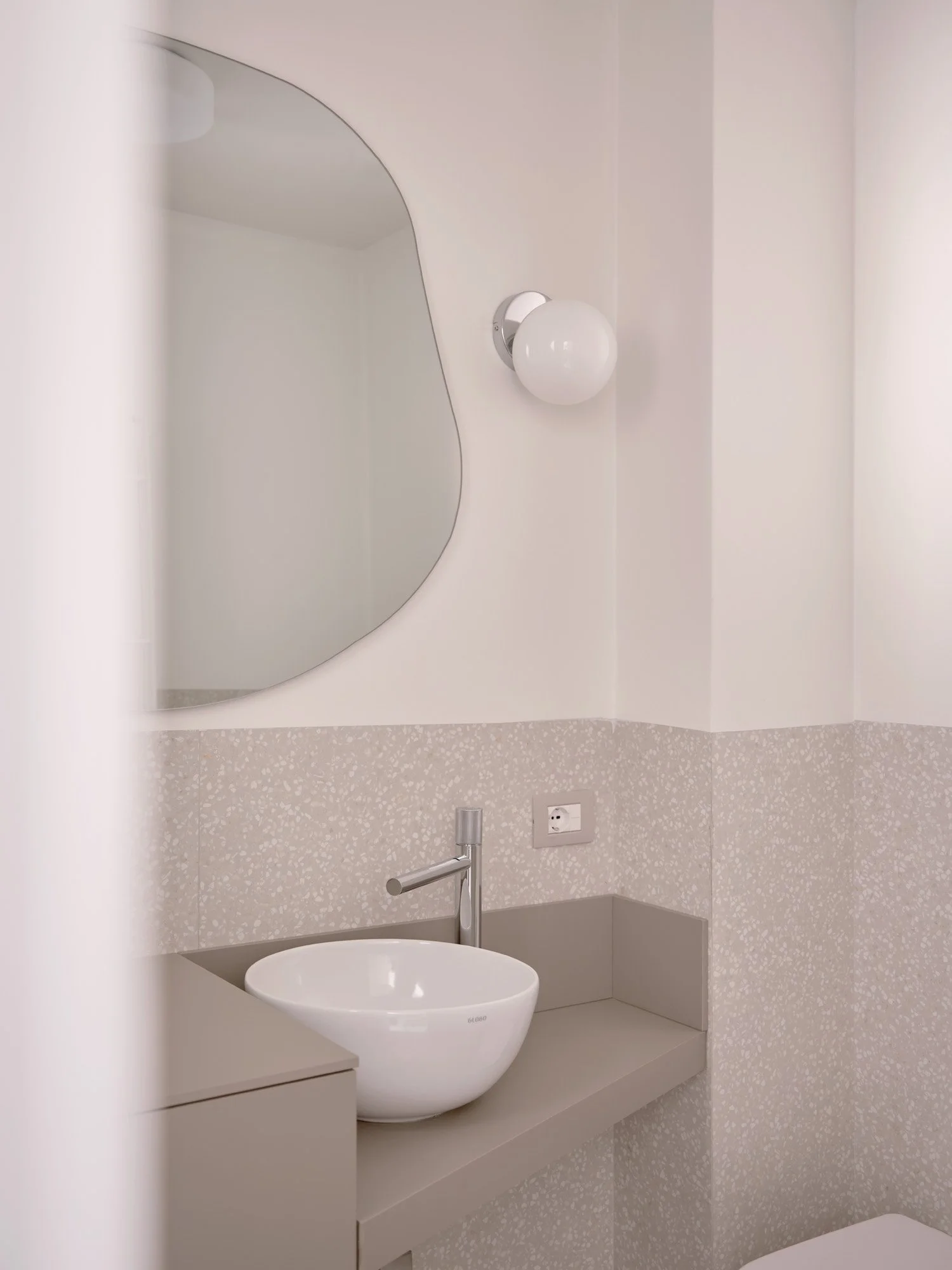 Specchio rotondo, lampada a sfera, lavabo bianco sopra un piano in vetro, presa di corrente, pareti bianche e rivestimento a mosaico chiaro.