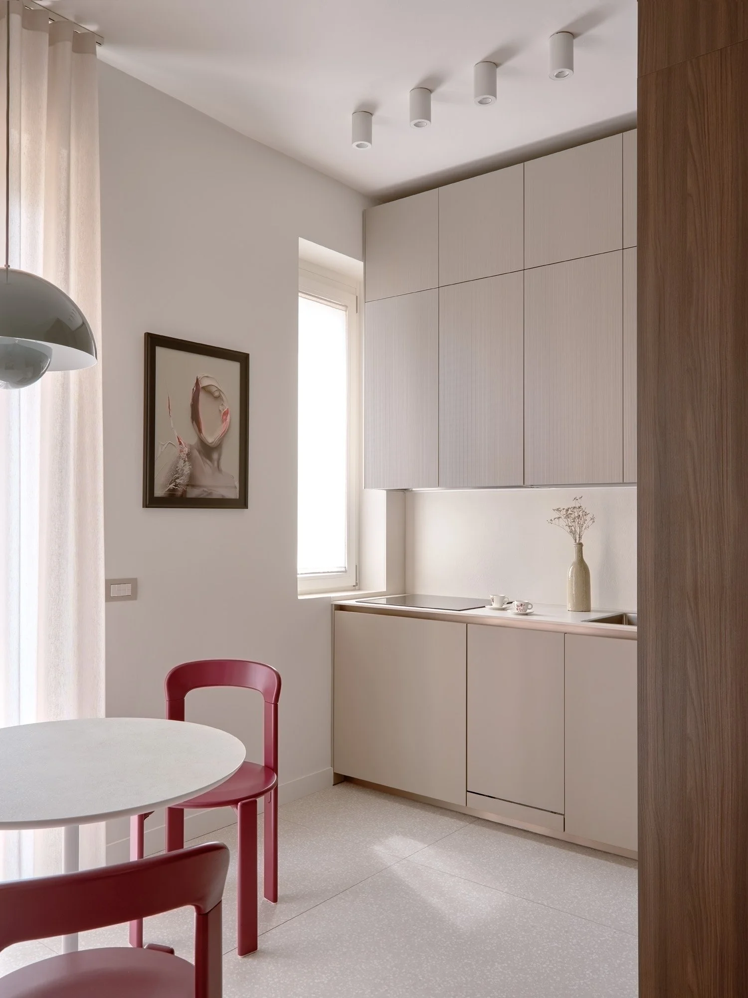 Cucina minimalista con pensili bianchi, finestra, occhielli di luce sul soffitto, parede decorativa con quadro e un vaso con fiori secchi sul piano cucina, sedia rossa e tavolo rotondo