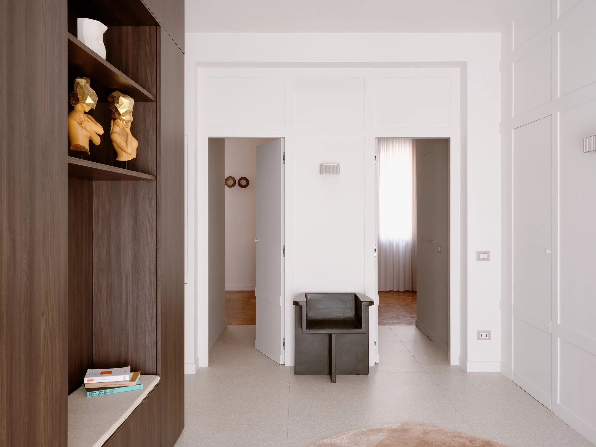 Ingresso con pareti bianche, una sedia nera e un armadio bianco. Sul lato sinistro, scaffale di legno con statue e libri.