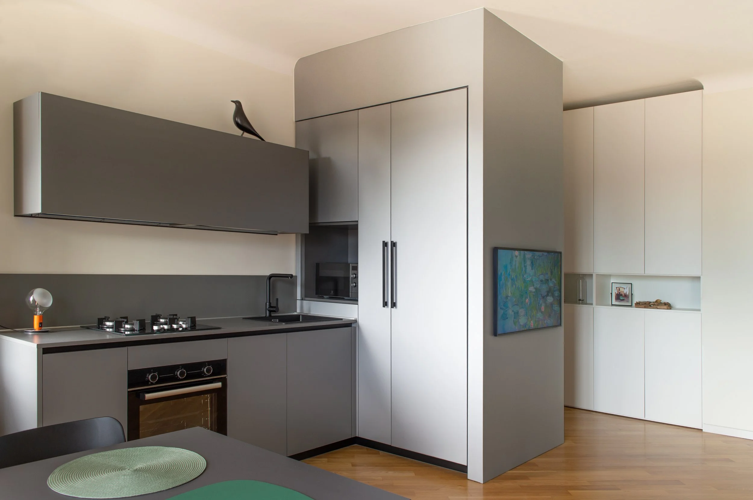Cucina moderna con mobili bianco e grigio, forno, piano cottura a gas, microonde, e tavolo con sedia, decorata con una scultura di uccello in alto