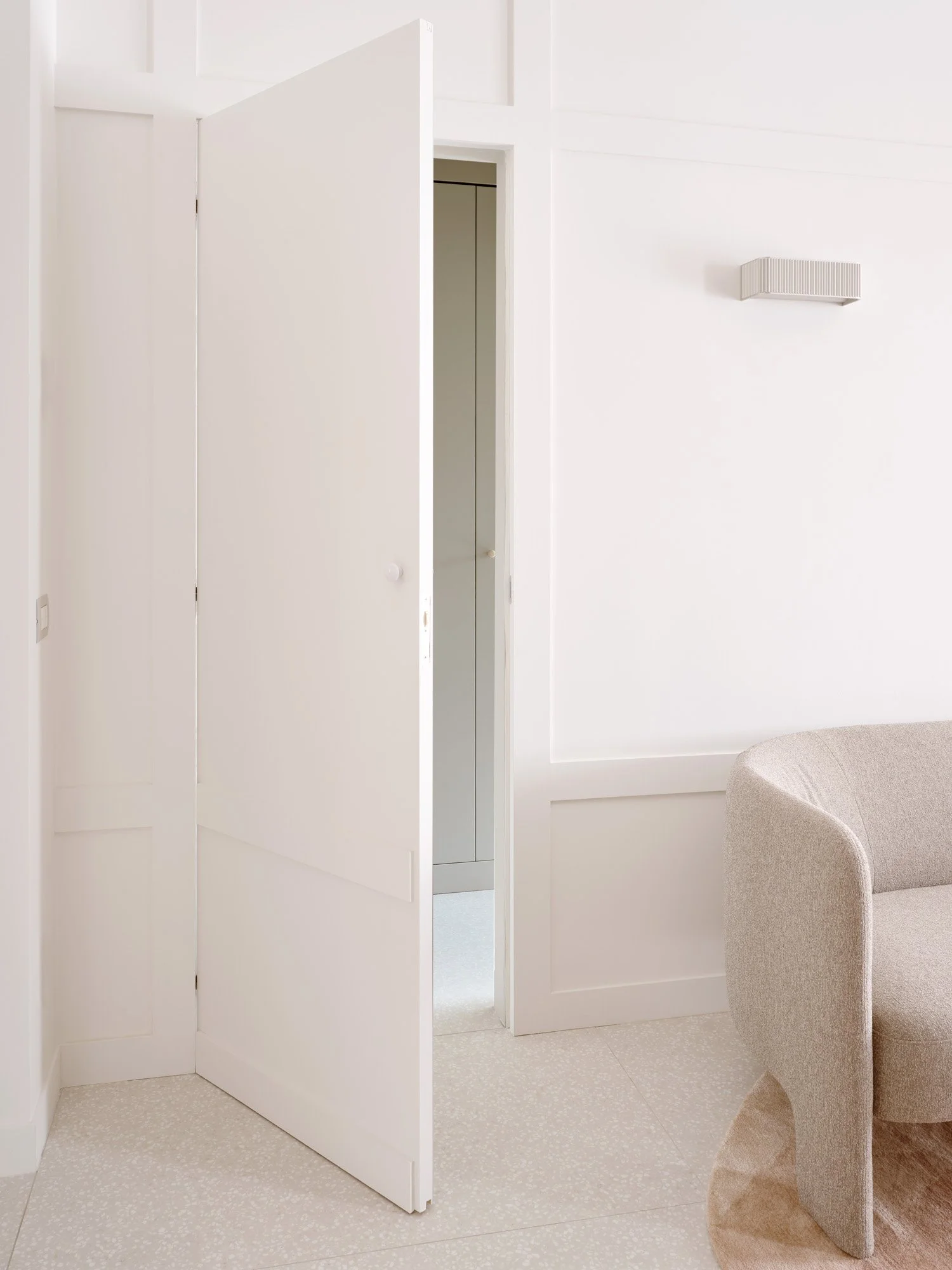 Interior di una stanza con pareti e porta bianche, divano beige e lampada a parete moderna.