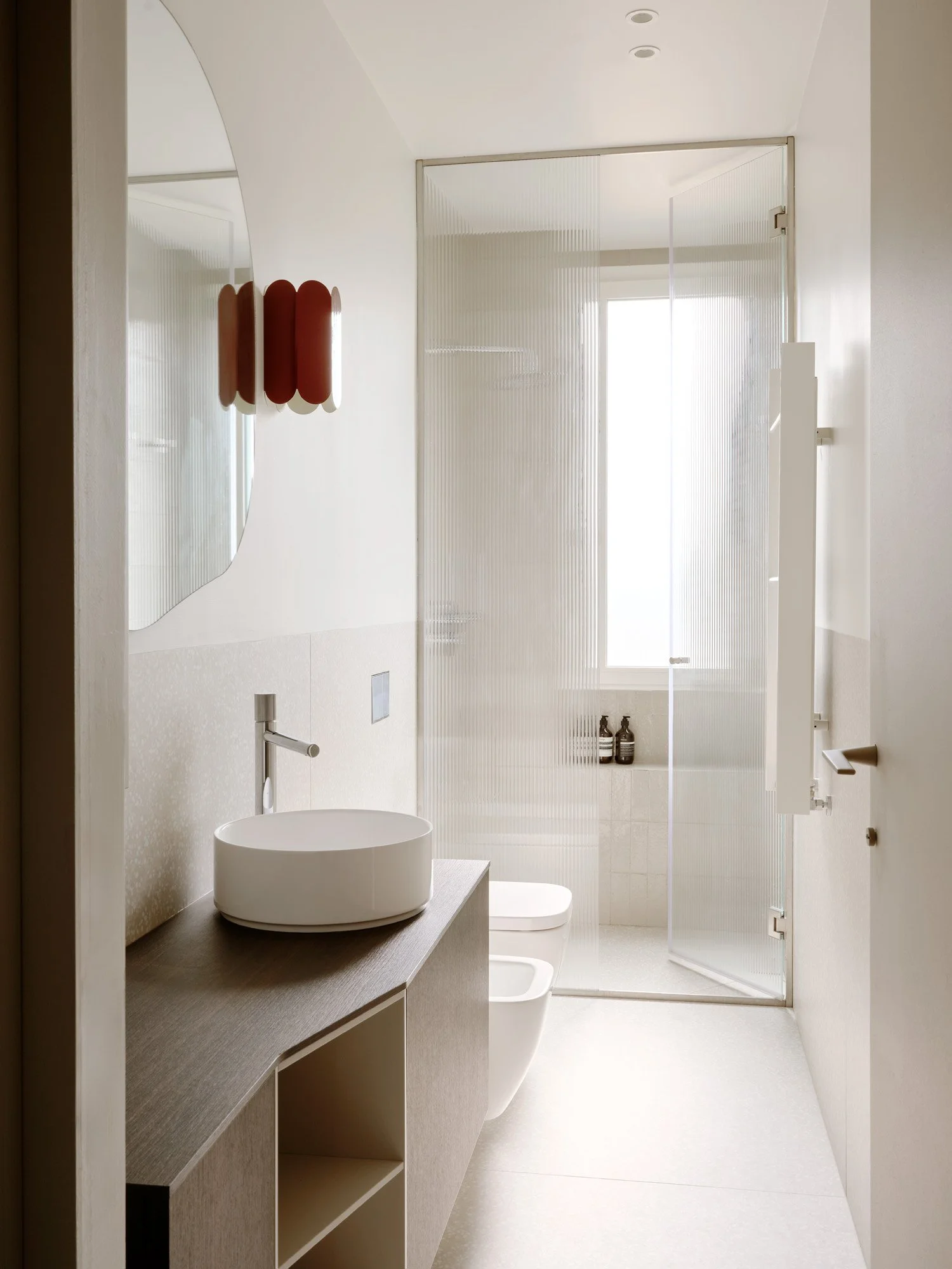 Bagno con lavabo rotondo bianco sopra un mobile in legno chiaro, specchio rotondo, lavabo e bidet bianchi, pareti e pavimento chiari, doccia con parete di vetro smerigliato e finestra con luce naturale, dettagli di arredamento minimalista.