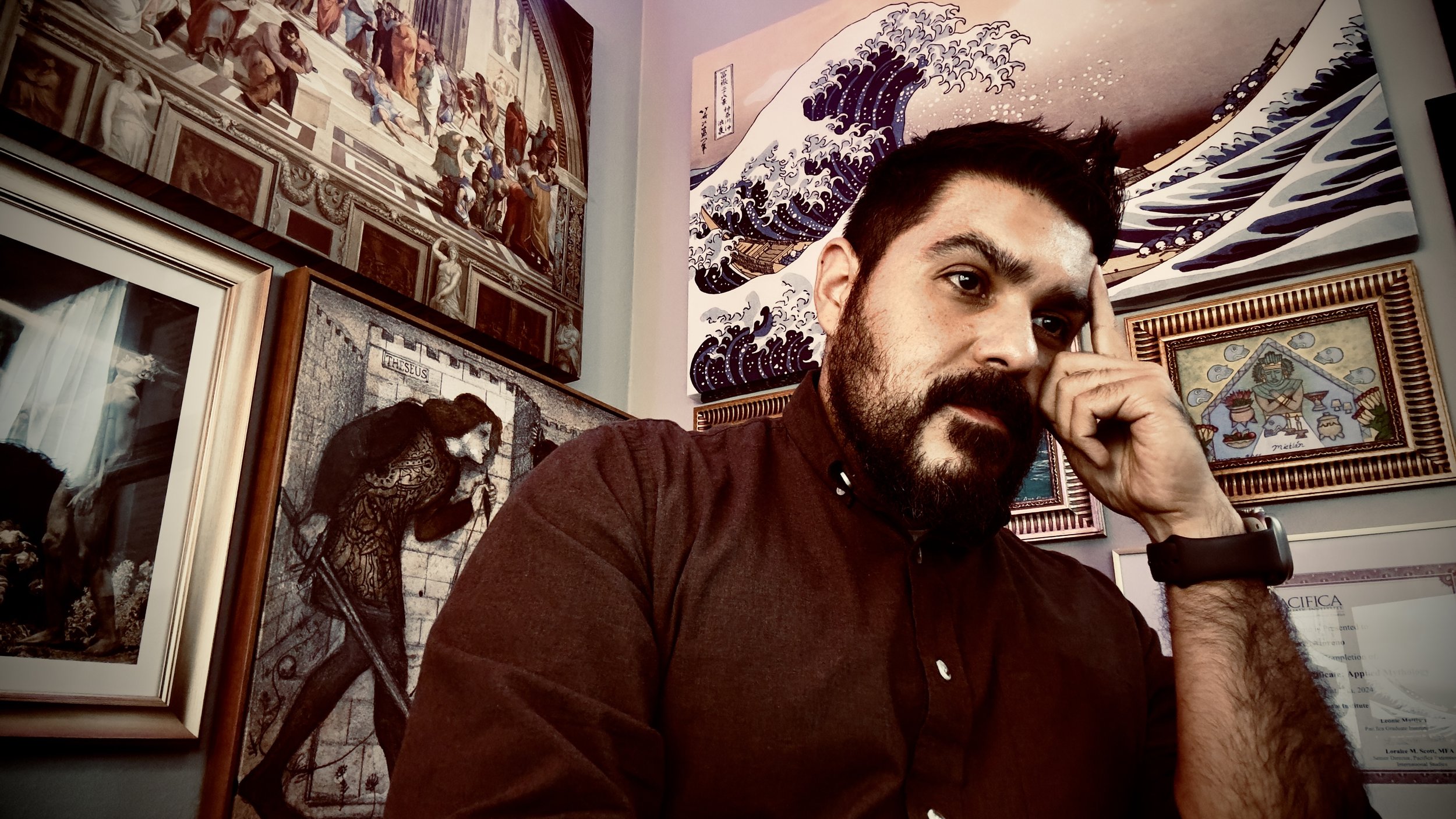 A man with a beard and dark hair, wearing a dark shirt and a black wristwatch, posing in front of a wall decorated with various framed artworks, including a painting of a wave, a mural of a medieval scene, and other colorful art pieces.