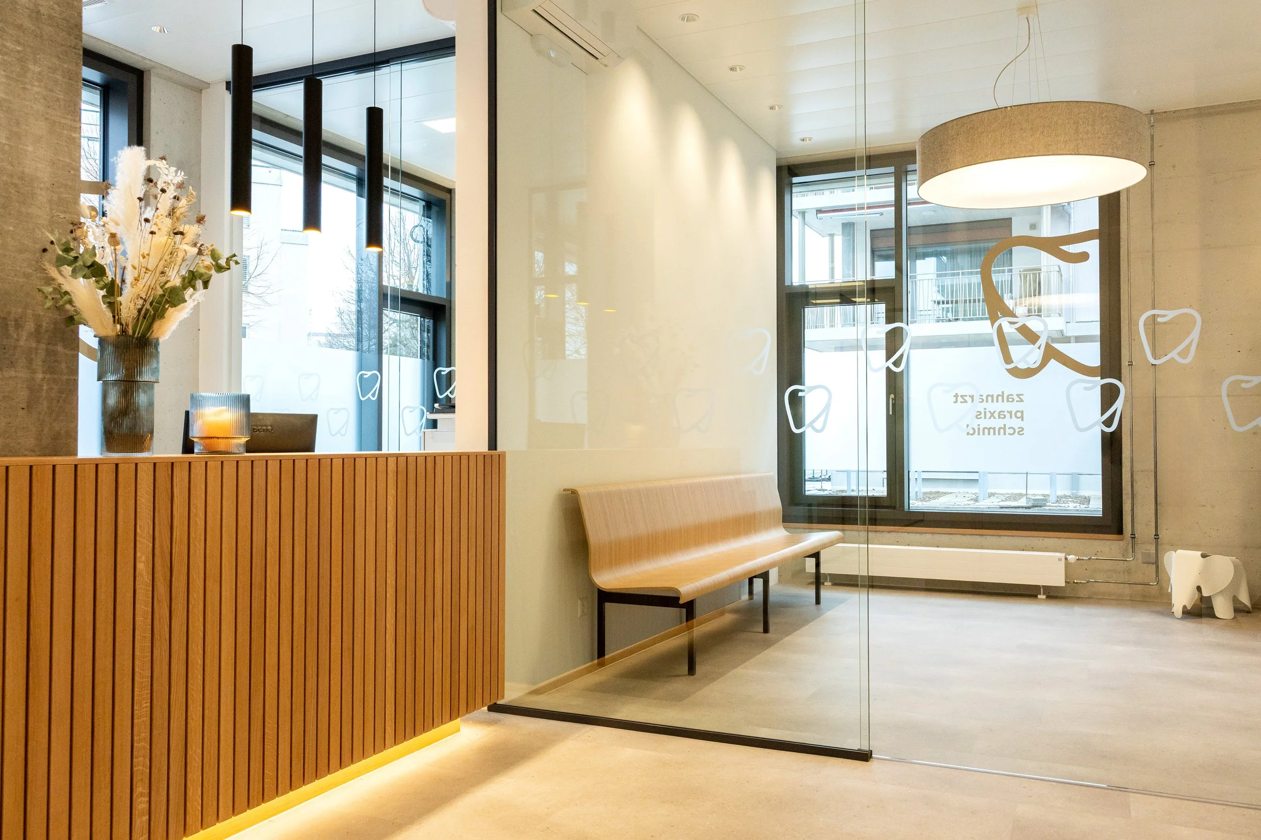 Moderner Wartebereich einer Praxis mit warmem Licht, hellen Holzelementen, mehreren Stuehlen und einer grossen Glaswand, die eine ruhige und einladende Atmosphaere schafft.