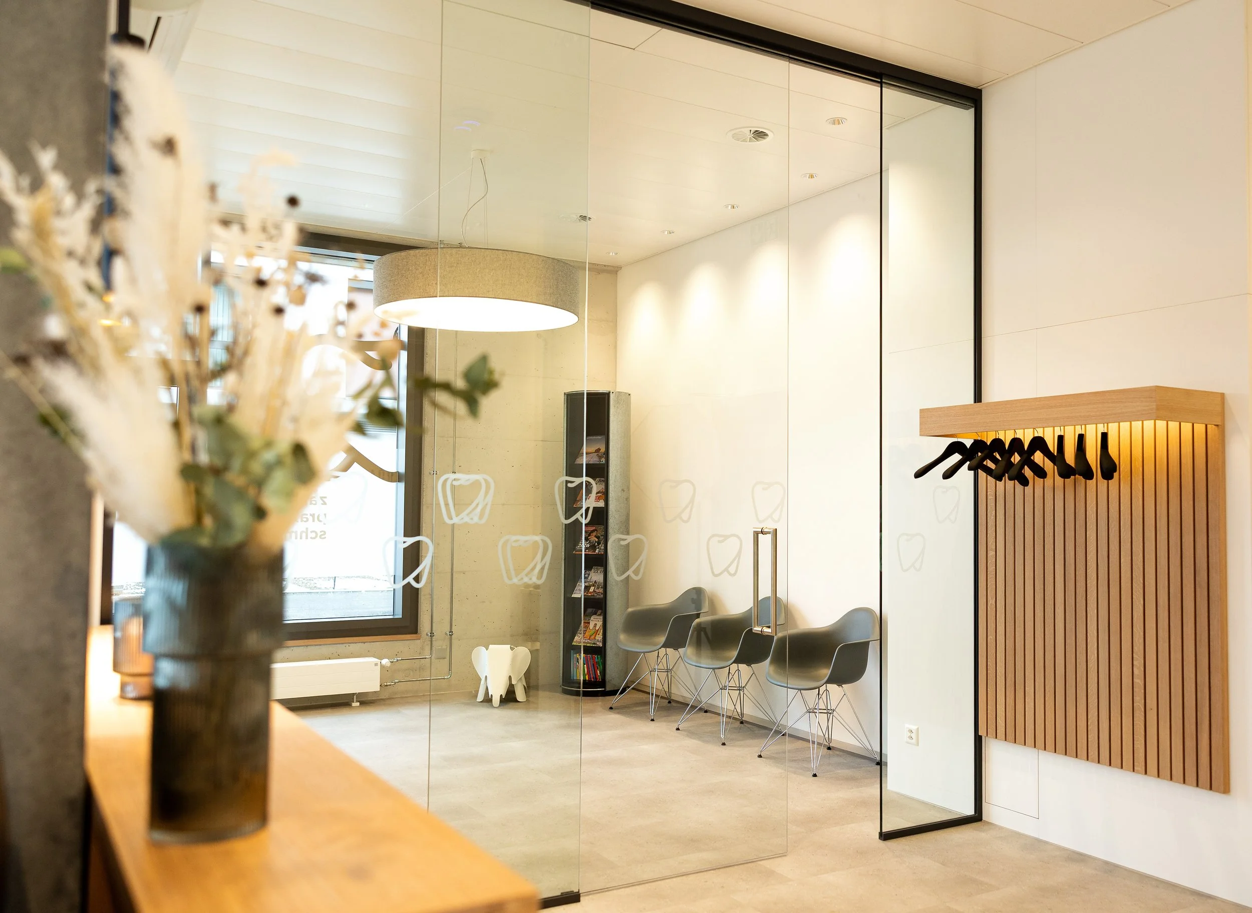 Helles Wartezimmer einer Praxis mit mehreren modernen Stühlen, einer großen Glaswand und warmem, einladendem Licht, das eine ruhige und freundliche Atmosphäre vermittelt.