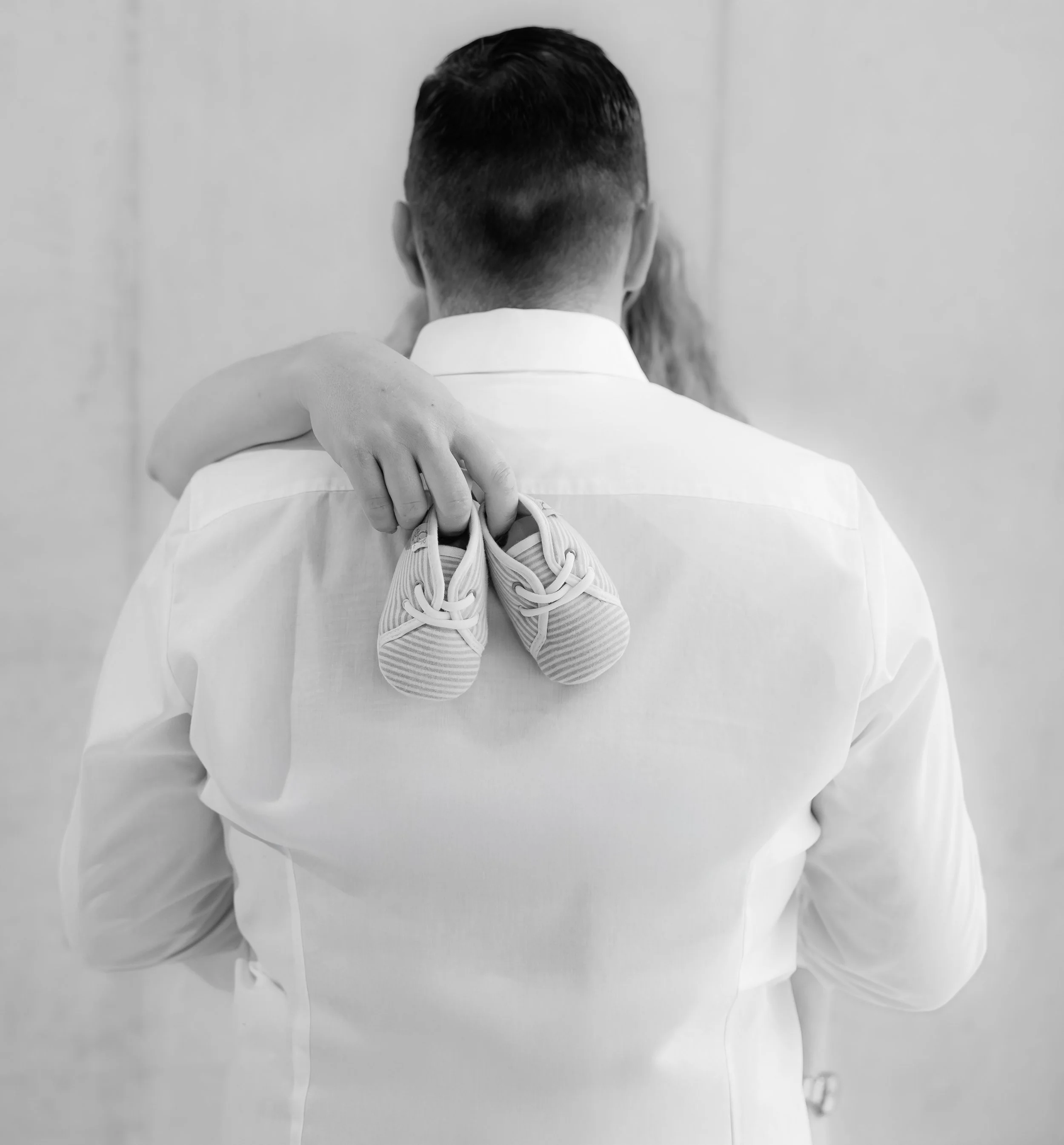 Schwarzweiss eines des Mannes von hinten, gekleidet in einem Hemd. Die Mutter hält von Hinten die Babyschuhe in der Hand.
