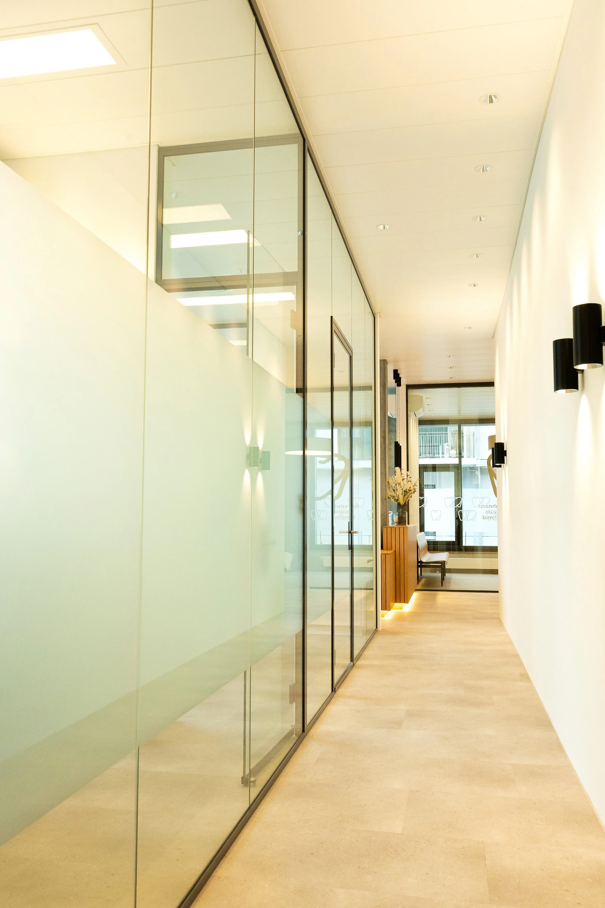 Lange Glaswand in einem hellen Praxisbereich mit warmem Licht, in dessen Mitte der Empfangsbereich sichtbar ist und eine ruhige, einladende Atmosphaere entsteht.