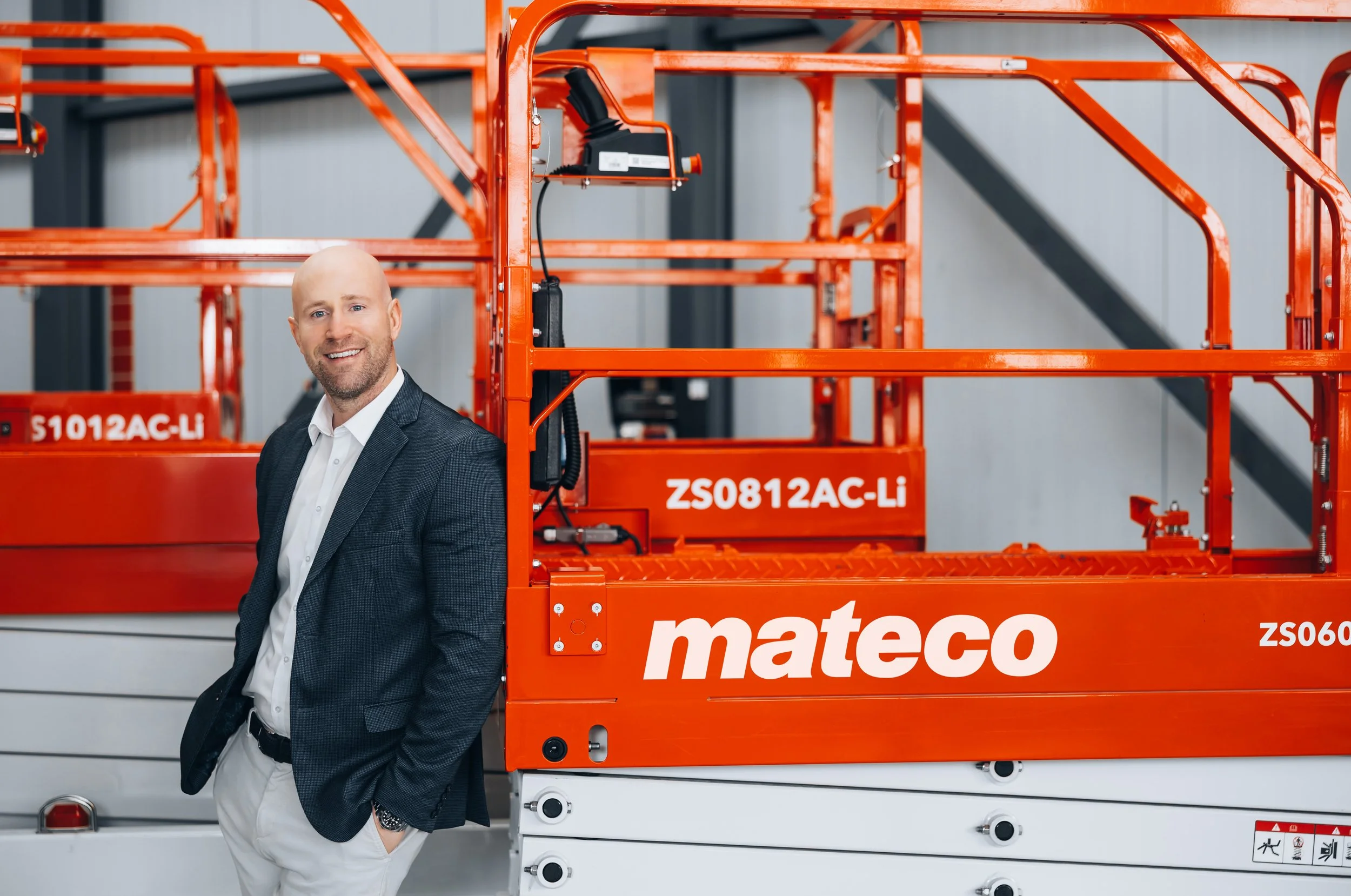 Ein Mann in einem dunklen Anzug lächelt und lehnt sich seitlich an eine rote Hebebühne.