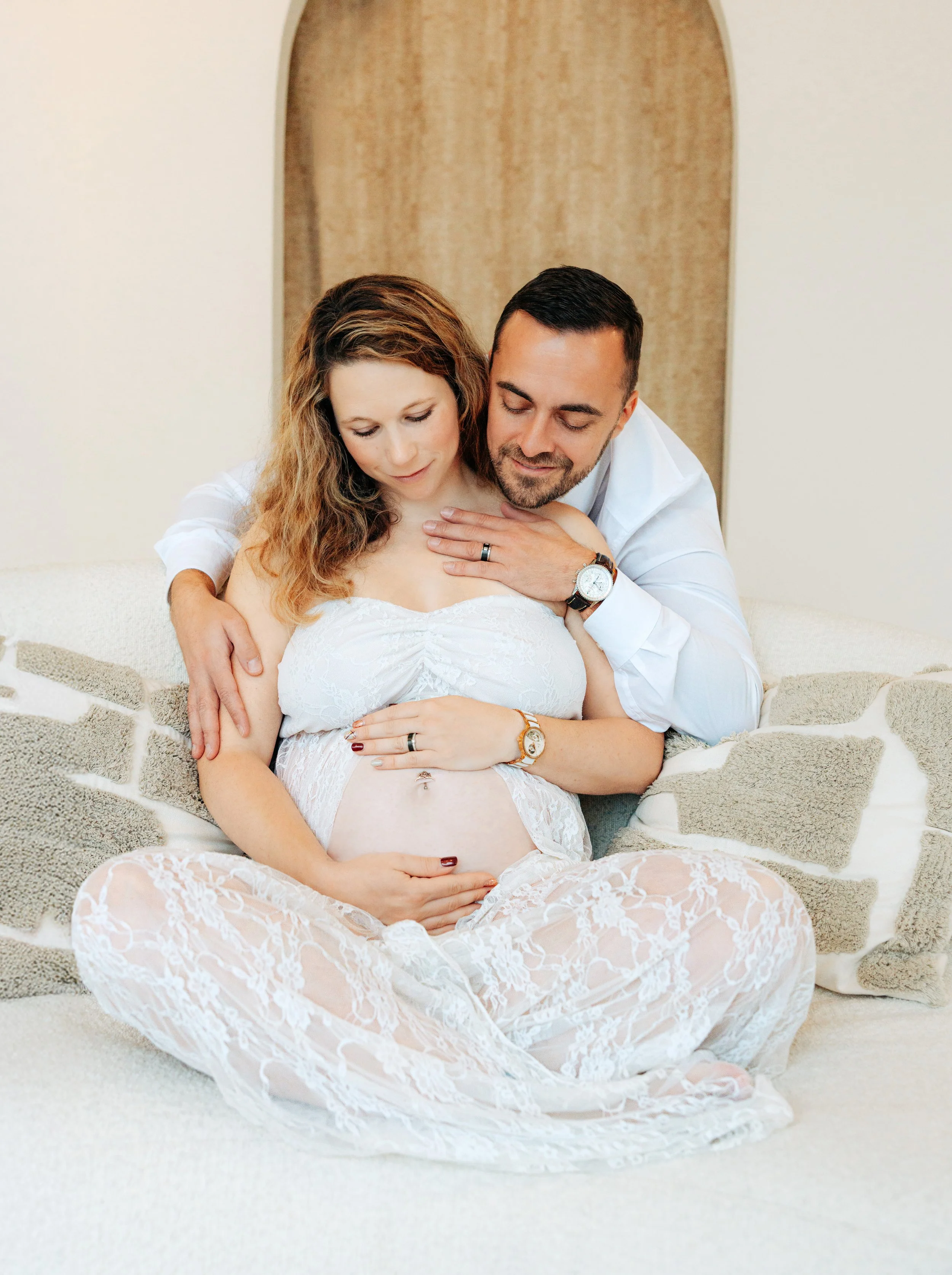 Die Eltern schauen beide lächelnd auf den Babybauch. Beide tragen weisse Kleidung.