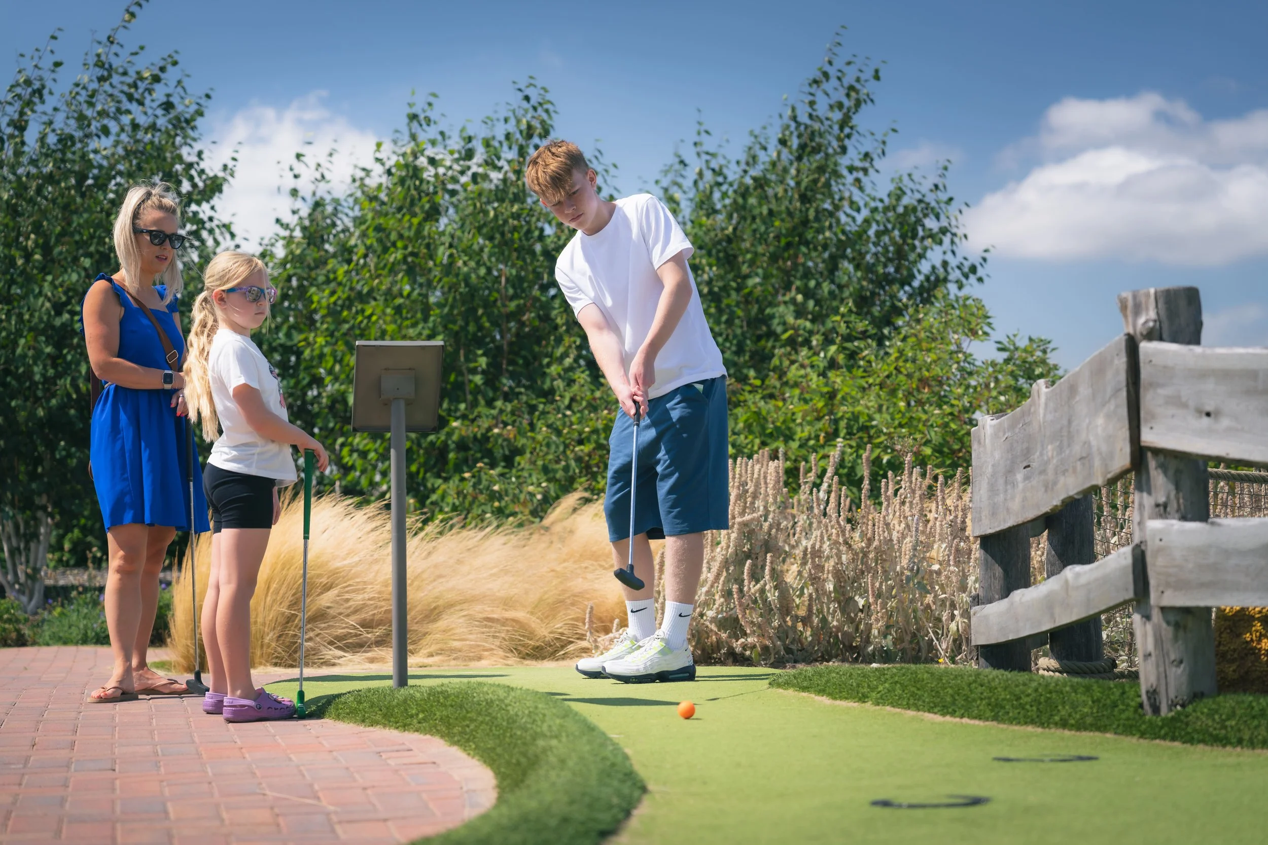 A young boy is playing mini golf on a green, with an orange golf ball nearby. Two girls and a woman are watching him; all are outdoors with trees and a blue sky in the background.