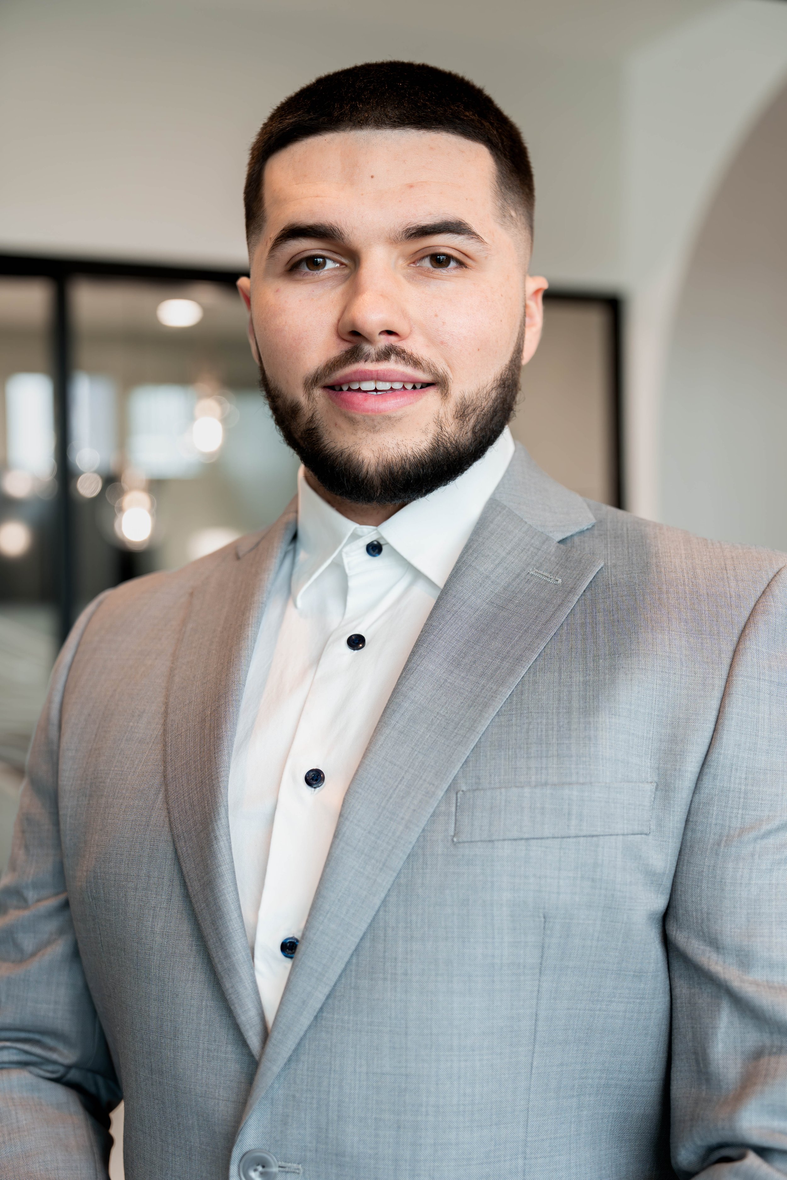 Man in a gray suit and white shirt smiling indoors with blurred background.