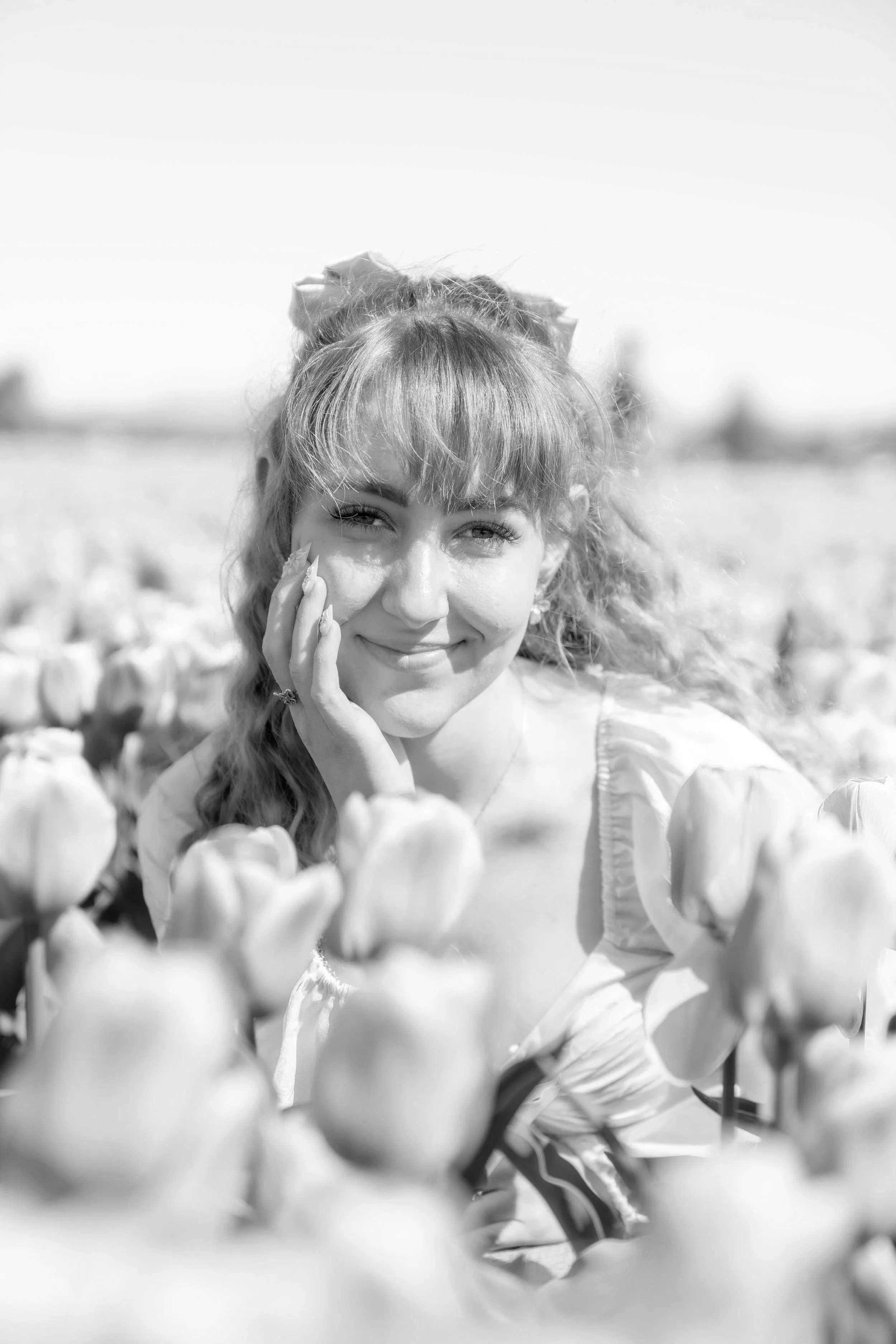 A smiling woman with wavy hair and bangs, standing in a field of tulips, touching her face gently.