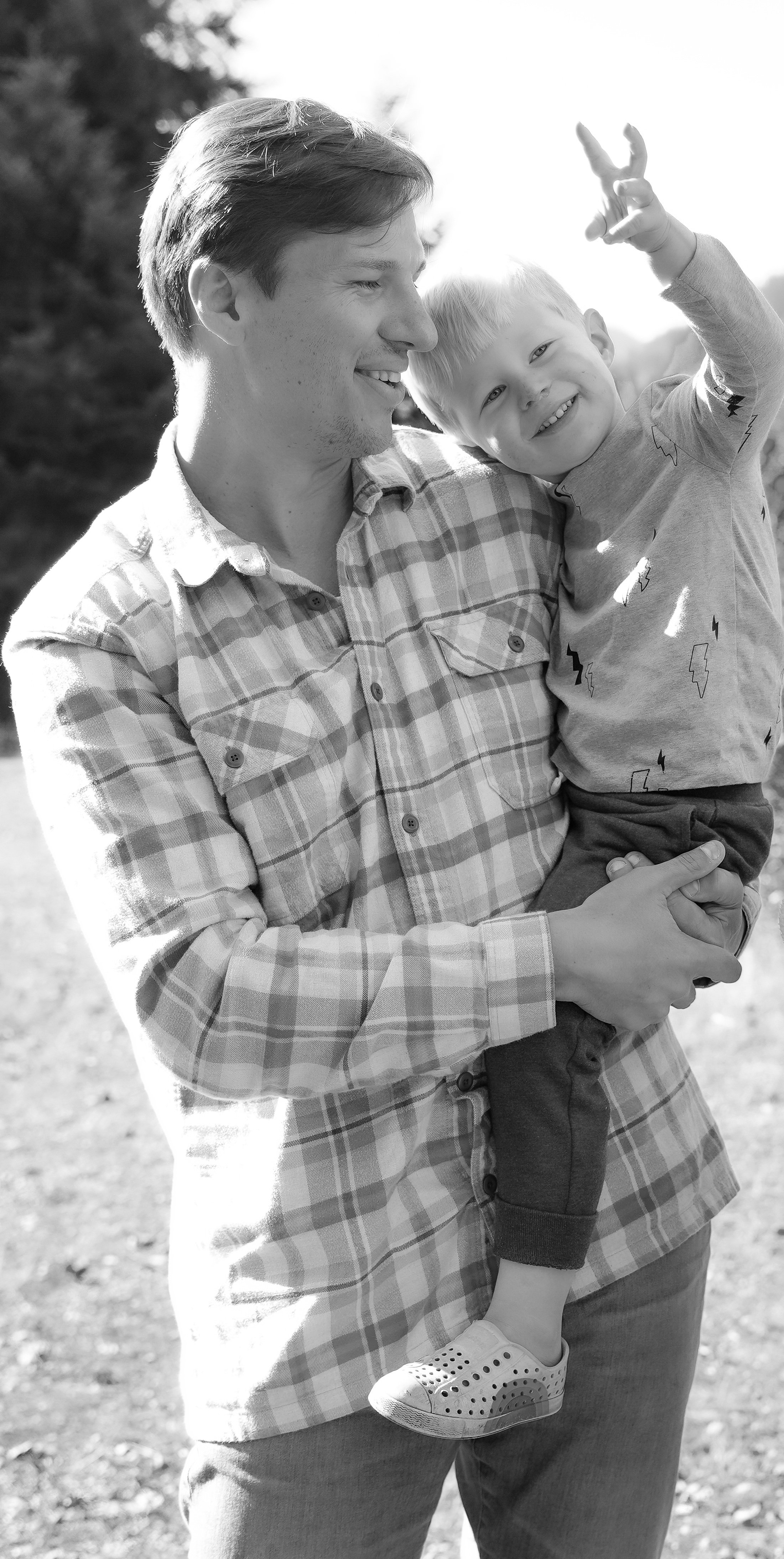 A man holding a smiling young boy in his arms outdoors, both facing each other, with the boy waving. The scene is bright, possibly in a park or natural setting.