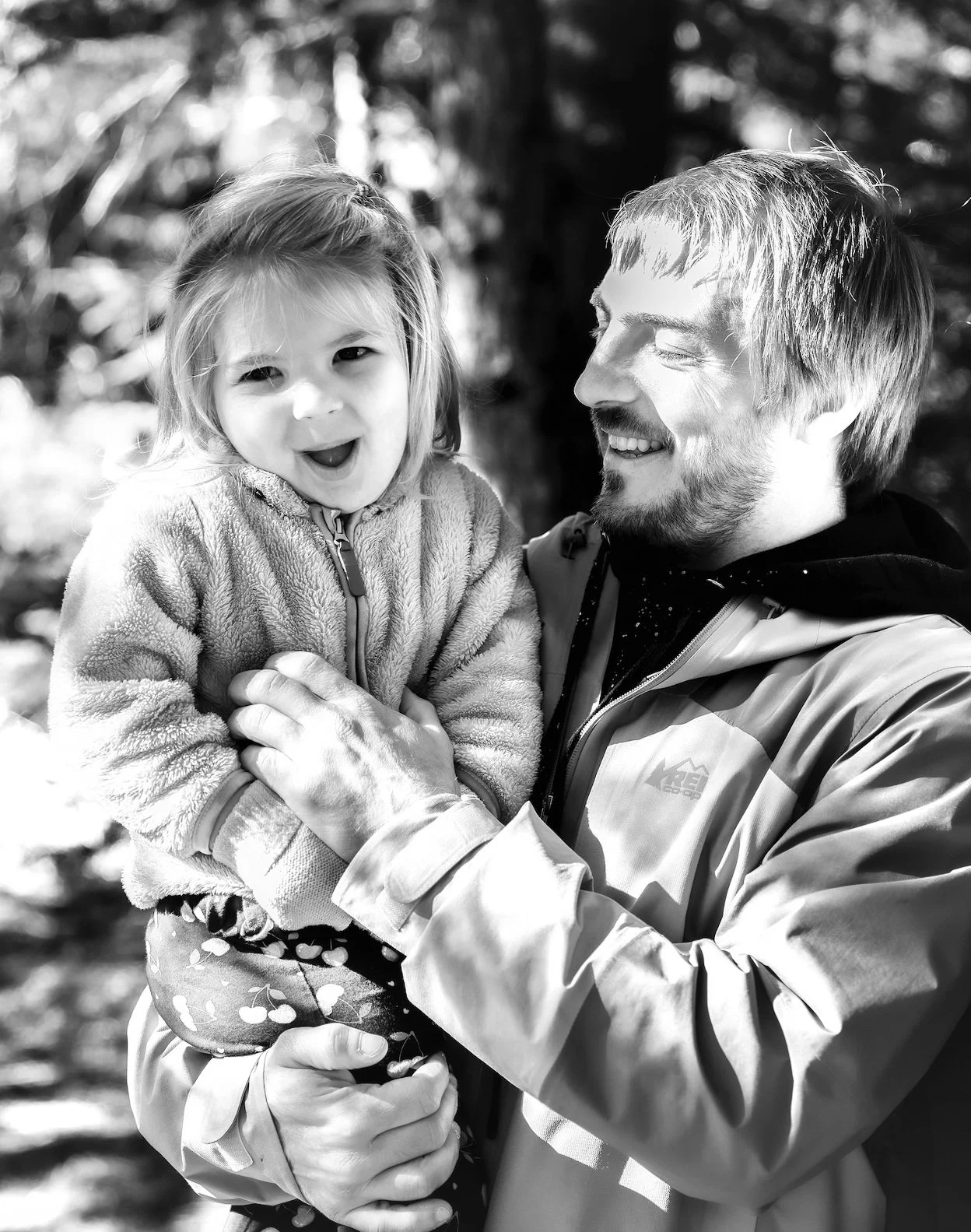 A man holding a young girl outdoors in a wooded area, both smiling and enjoying the moment, in black and white.