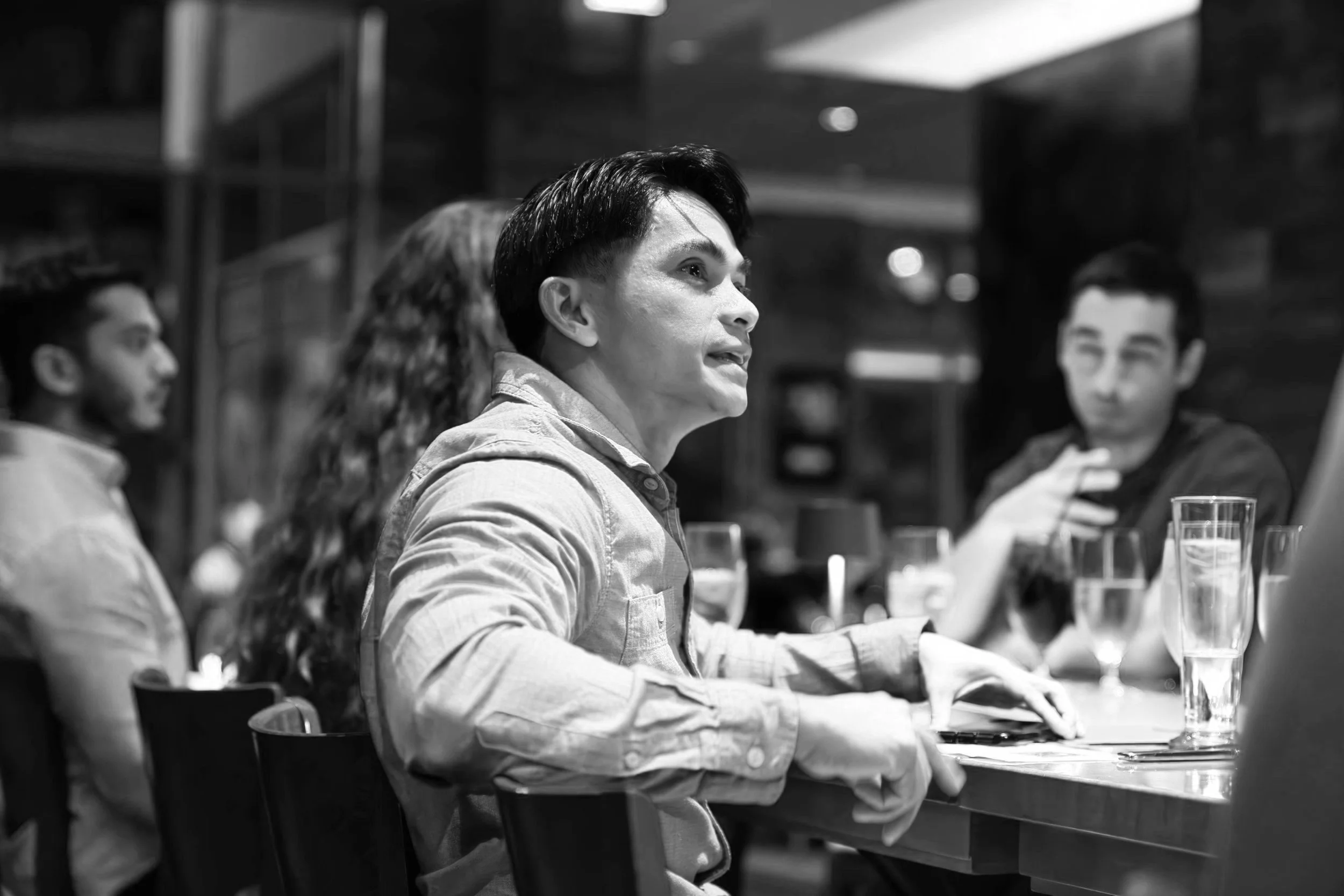 Group of people sitting at a table in a restaurant or cafe, engaged in conversation, in black and white photo.