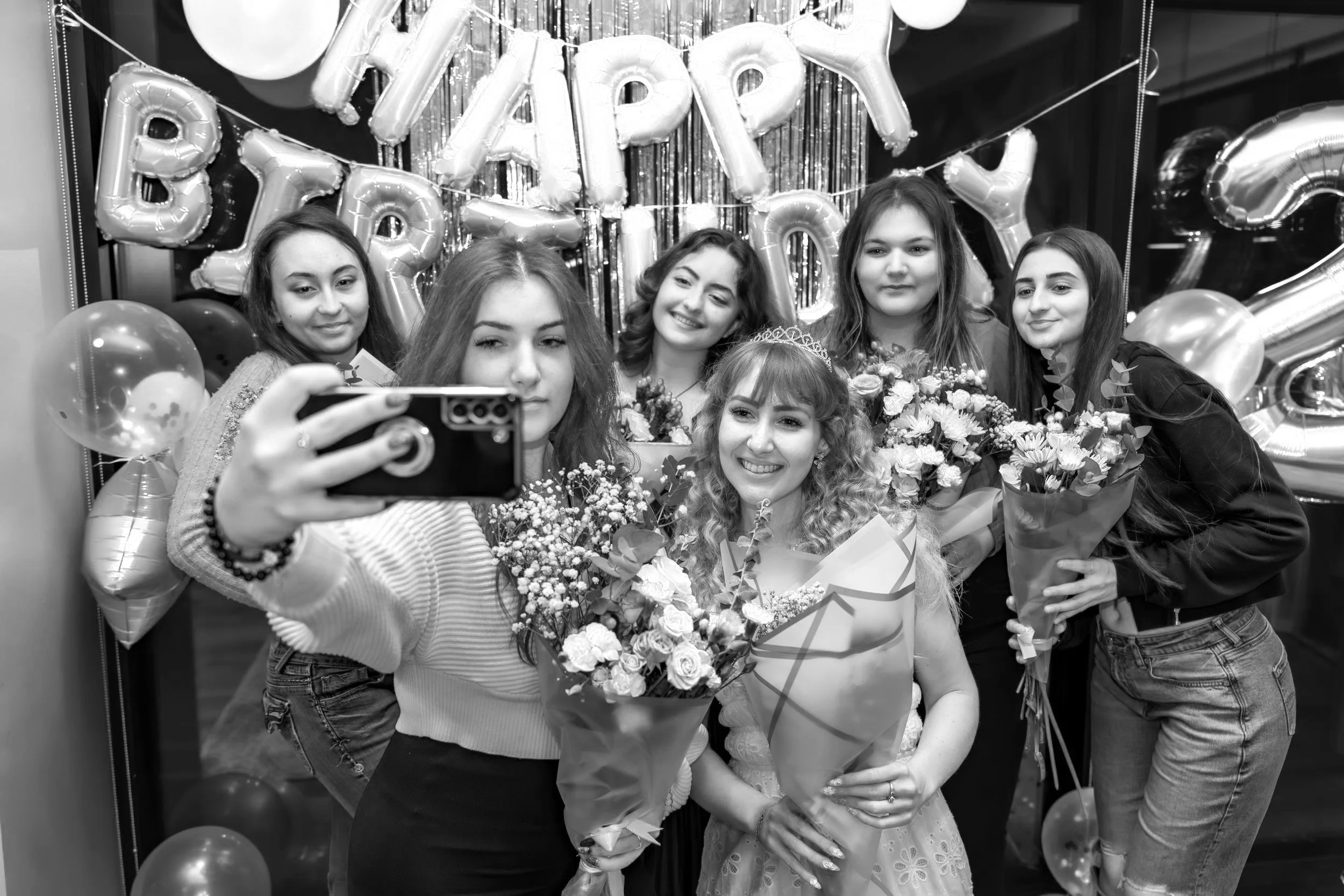 A group of six young women celebrating a birthday, with balloons and a 'Happy Birthday' banner in the background. One woman in the center is wearing a tiara and holding a bouquet of flowers, smiling for a selfie.