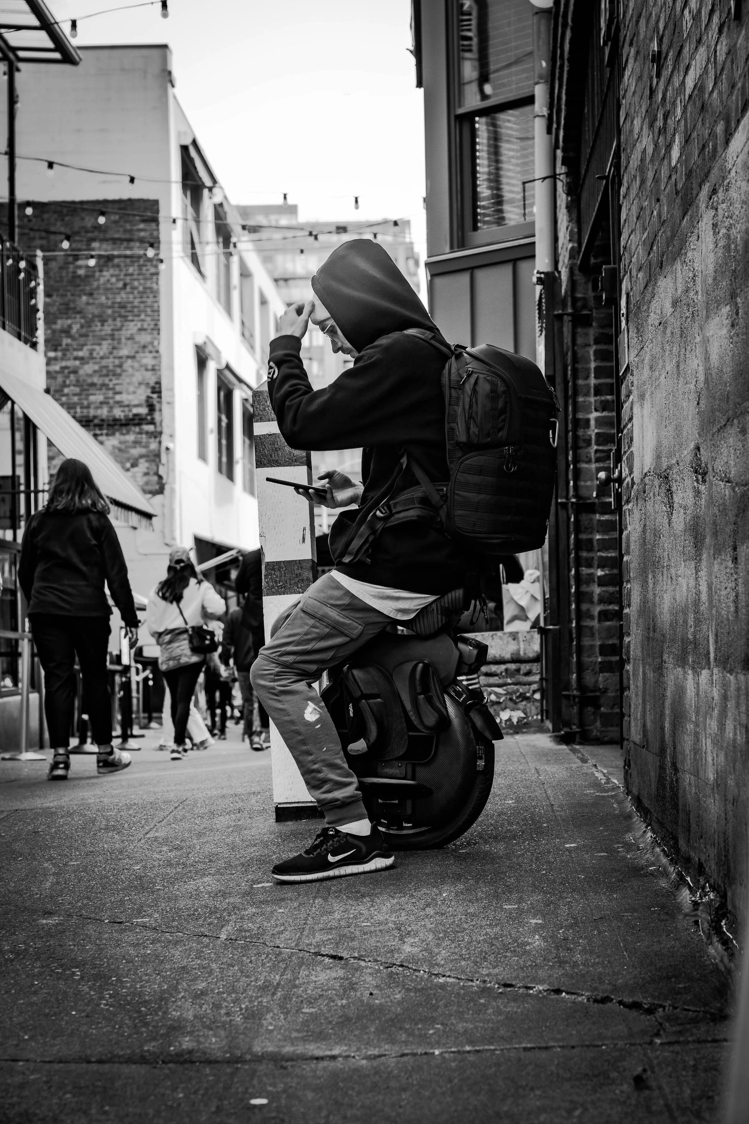 A person sitting on a unicycle on a city sidewalk, wearing a hoodie and backpack, looking at their phone. Several pedestrians are walking in the background.