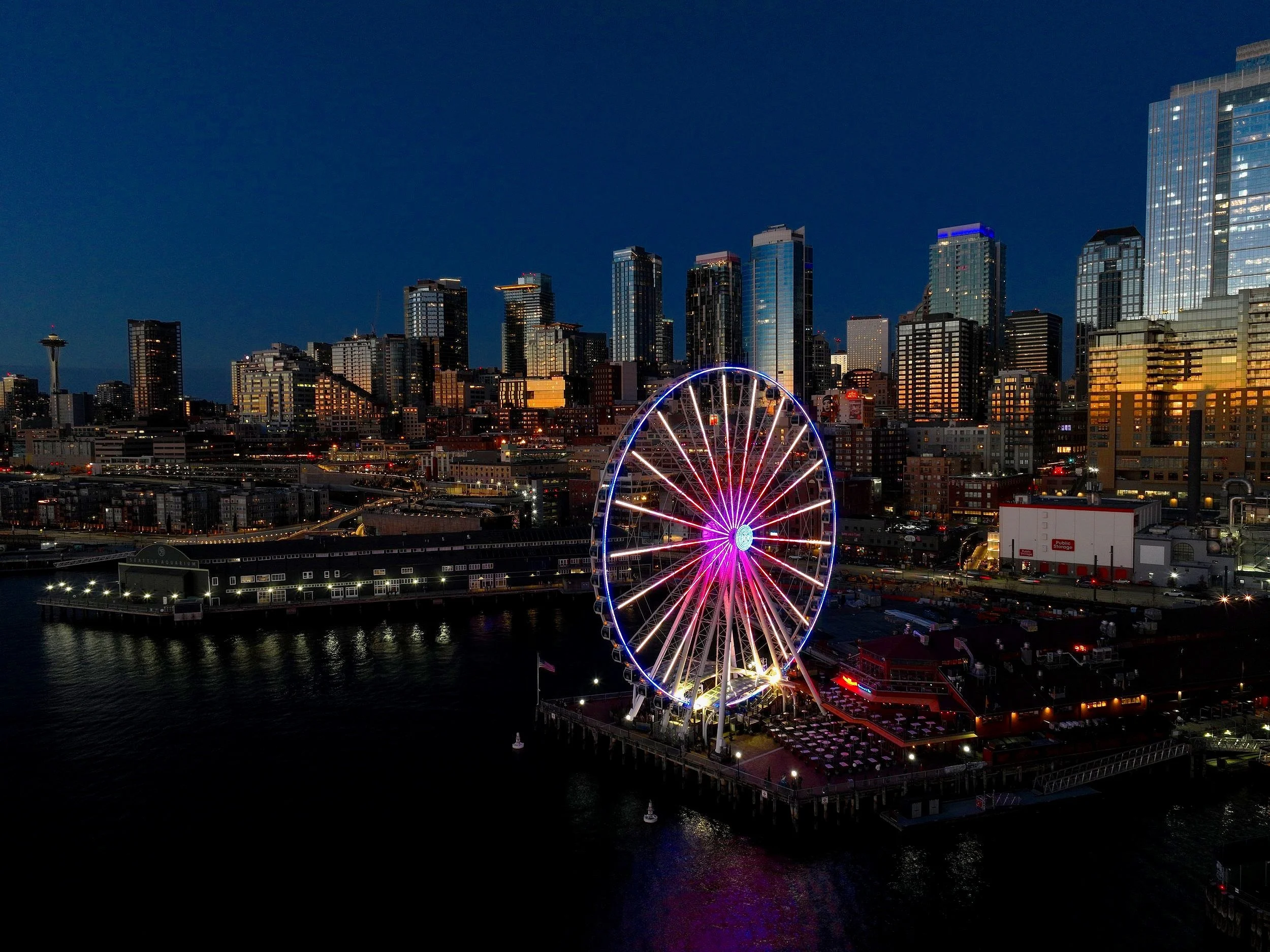 Nighttime cityscape of Seattle with illuminated Ferris wheel on the waterfront, tall skyscrapers including the Space Needle in the background.