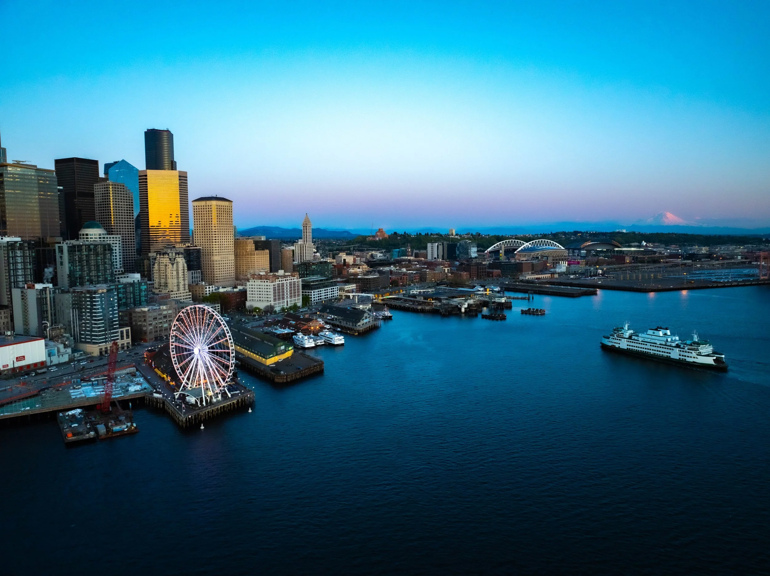 Seattle skyline at dusk with illuminated Ferris wheel, boats in the harbor, and the city buildings against a clear sky with Mount Rainier in the background.