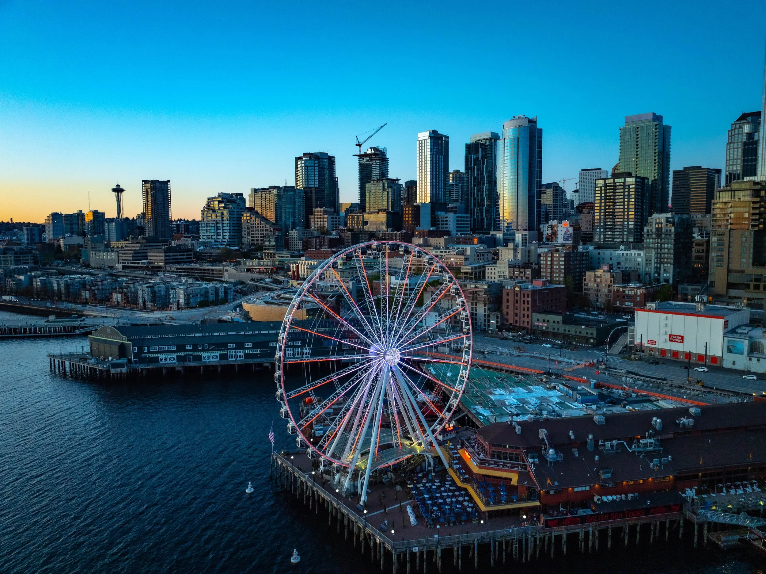 A city skyline at dusk featuring a large Ferris wheel along a waterfront pier in Seattle, with the Space Needle visible in the background and high-rise buildings illuminated by sunset.