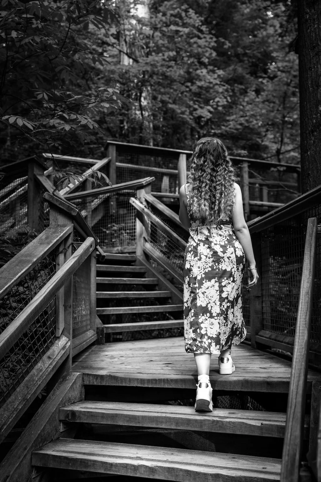 A woman with long curly hair wearing a floral dress and sneakers, walking up wooden stairs in a forest.