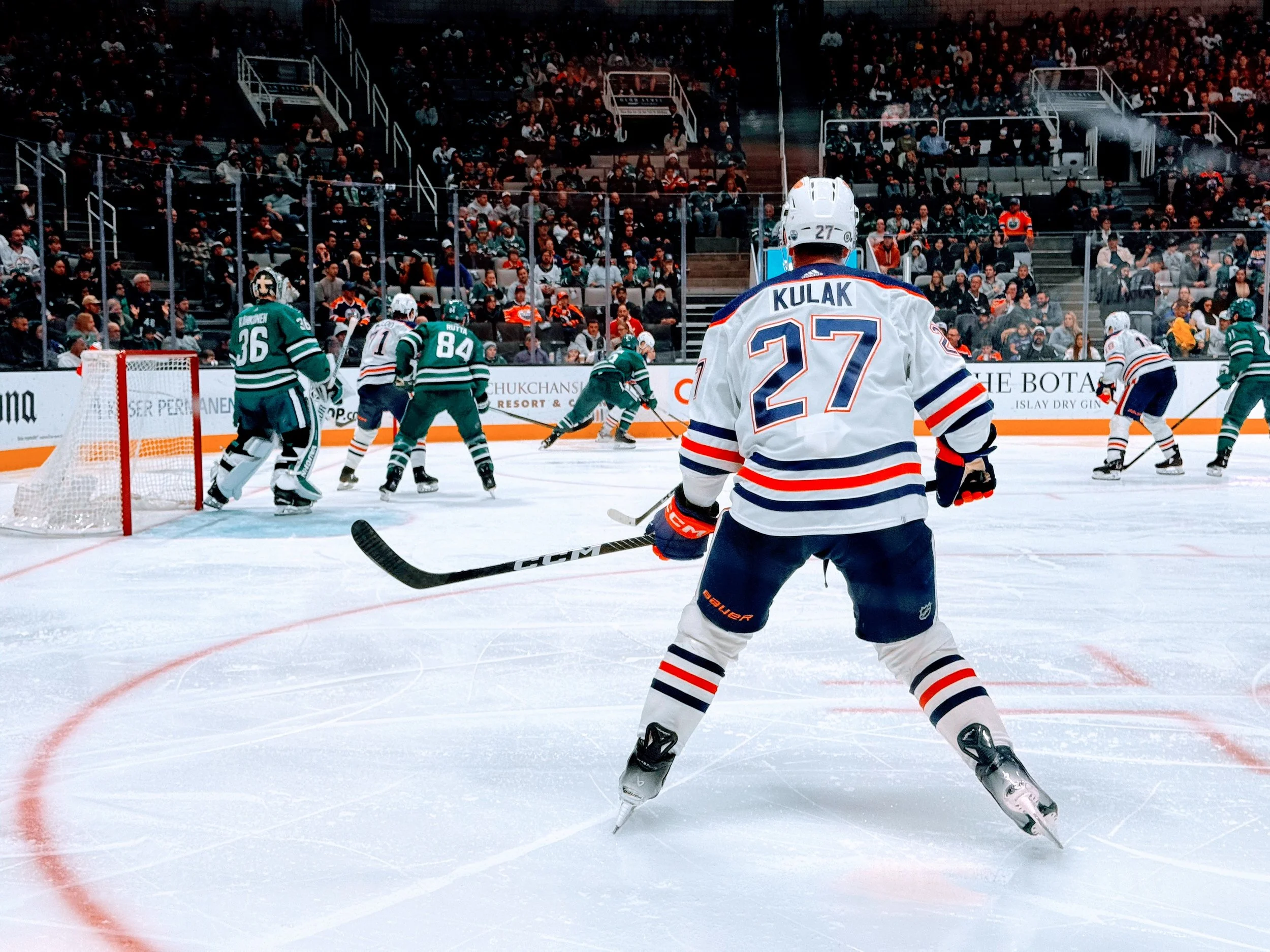 Hockey players on the ice during a game. A player with jersey number 27 stands in the foreground, with others near the goal and around the ice, and spectators in the stands.