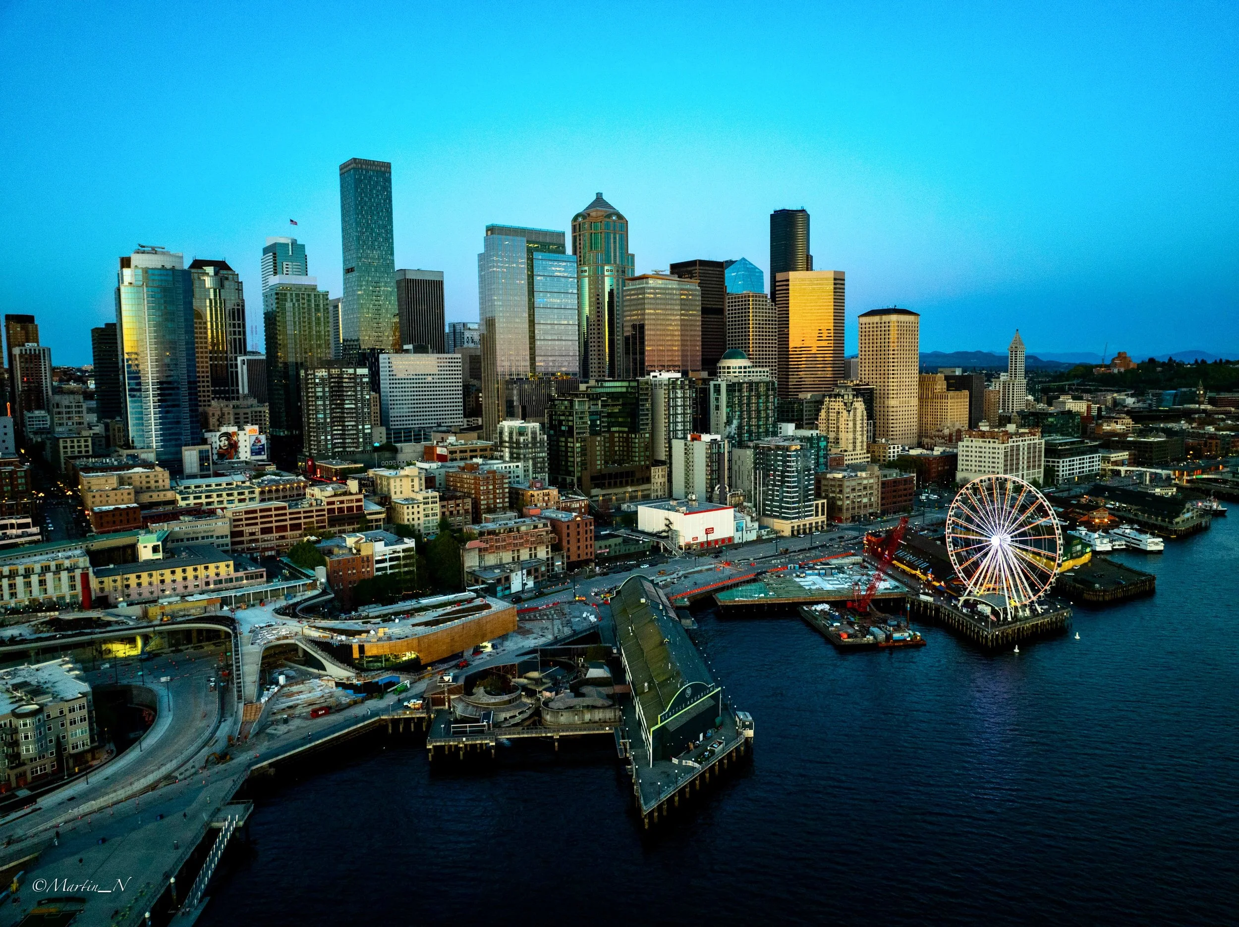 Aerial view of Seattle's downtown skyline at dusk, with tall buildings, a Ferris wheel by the waterfront, and a mix of water and city infrastructure.
