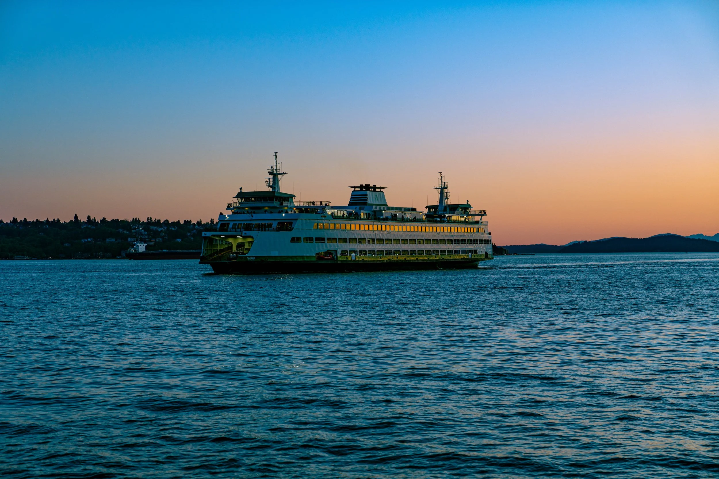 A large ferry boat sailing on calm water during sunset with a hilly shoreline in the background.