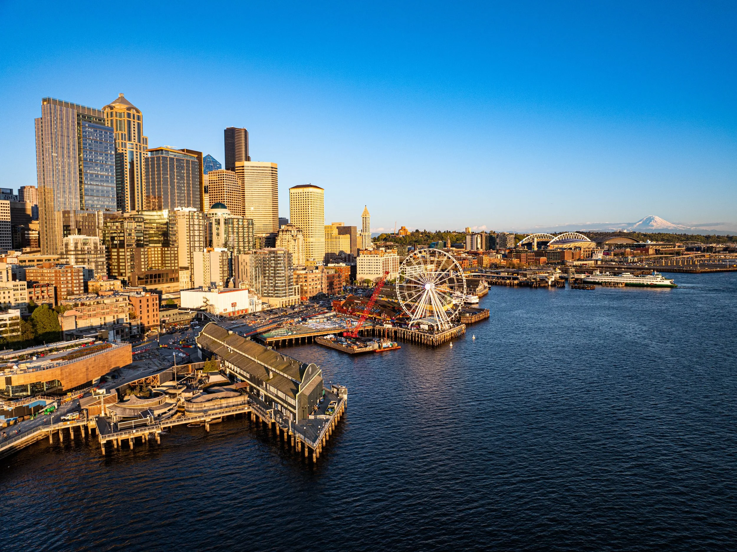 Aerial view of Seattle's skyline with tall modern buildings, a waterfront with a Ferris wheel, and Mount Rainier in the background, during daytime with clear blue sky.