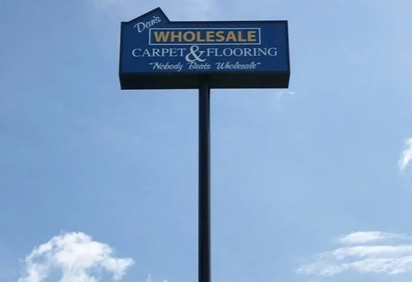 Sign for a business named "Dane's Wholesale Carpet & Flooring" with the slogan "Nobody Beats Wholesale" against a blue sky.