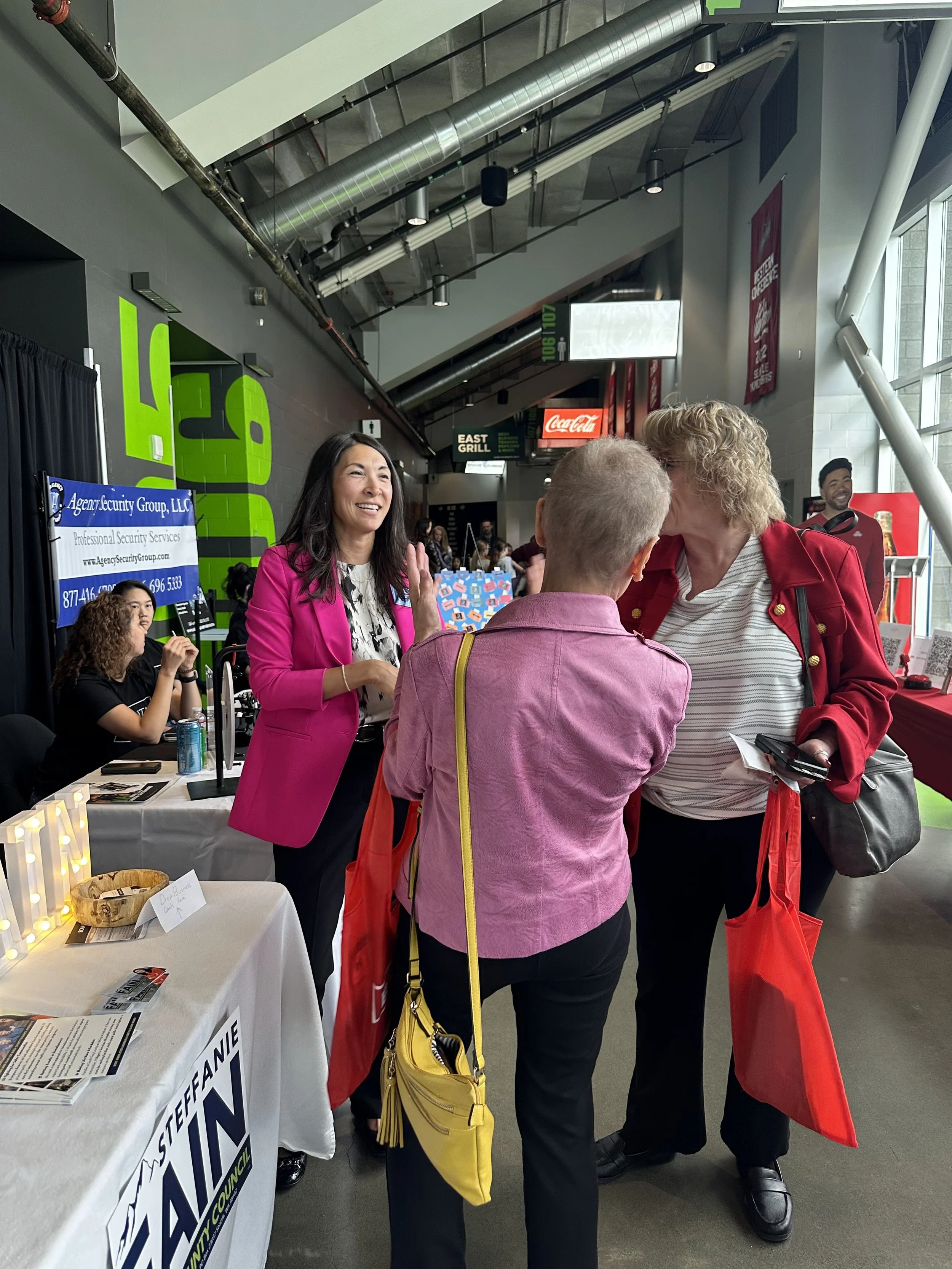 Three women engaged in conversation at an indoor event, with one woman wearing a pink blazer and another with blonde hair and a red jacket. There is a booth with a sign for Stephanie Hahn at a table, and a digital screen with company information in t