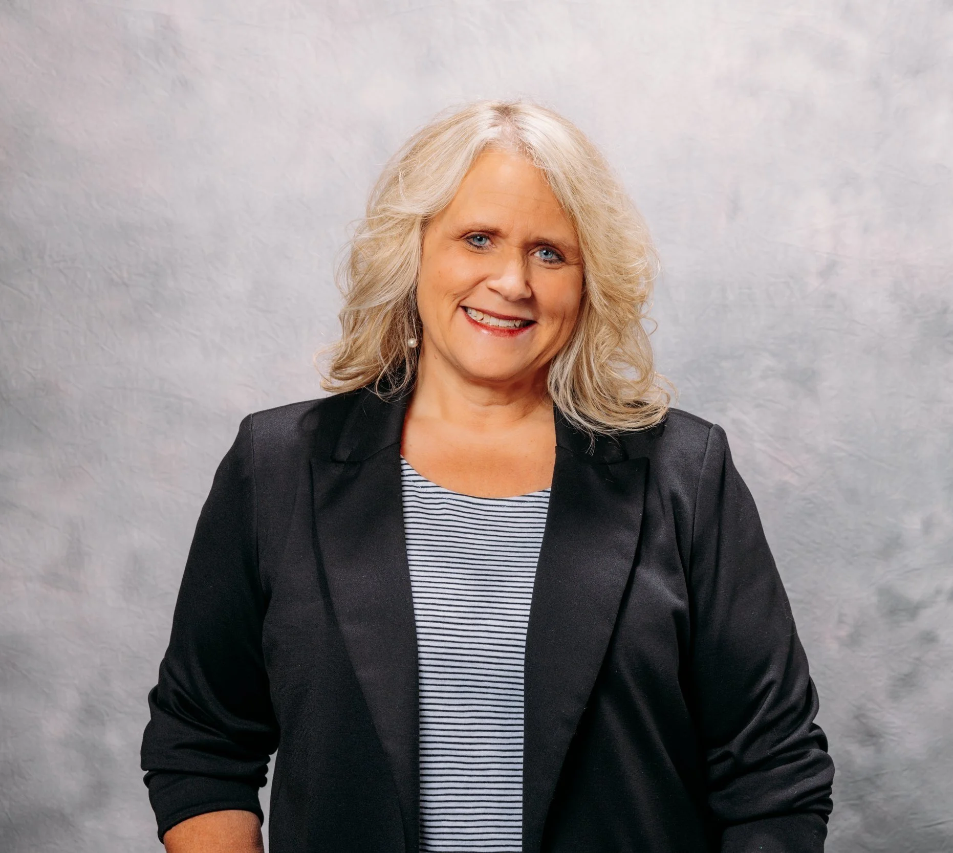 A woman with blonde, wavy hair smiling in front of a light-colored textured background, wearing a black blazer over a striped shirt.