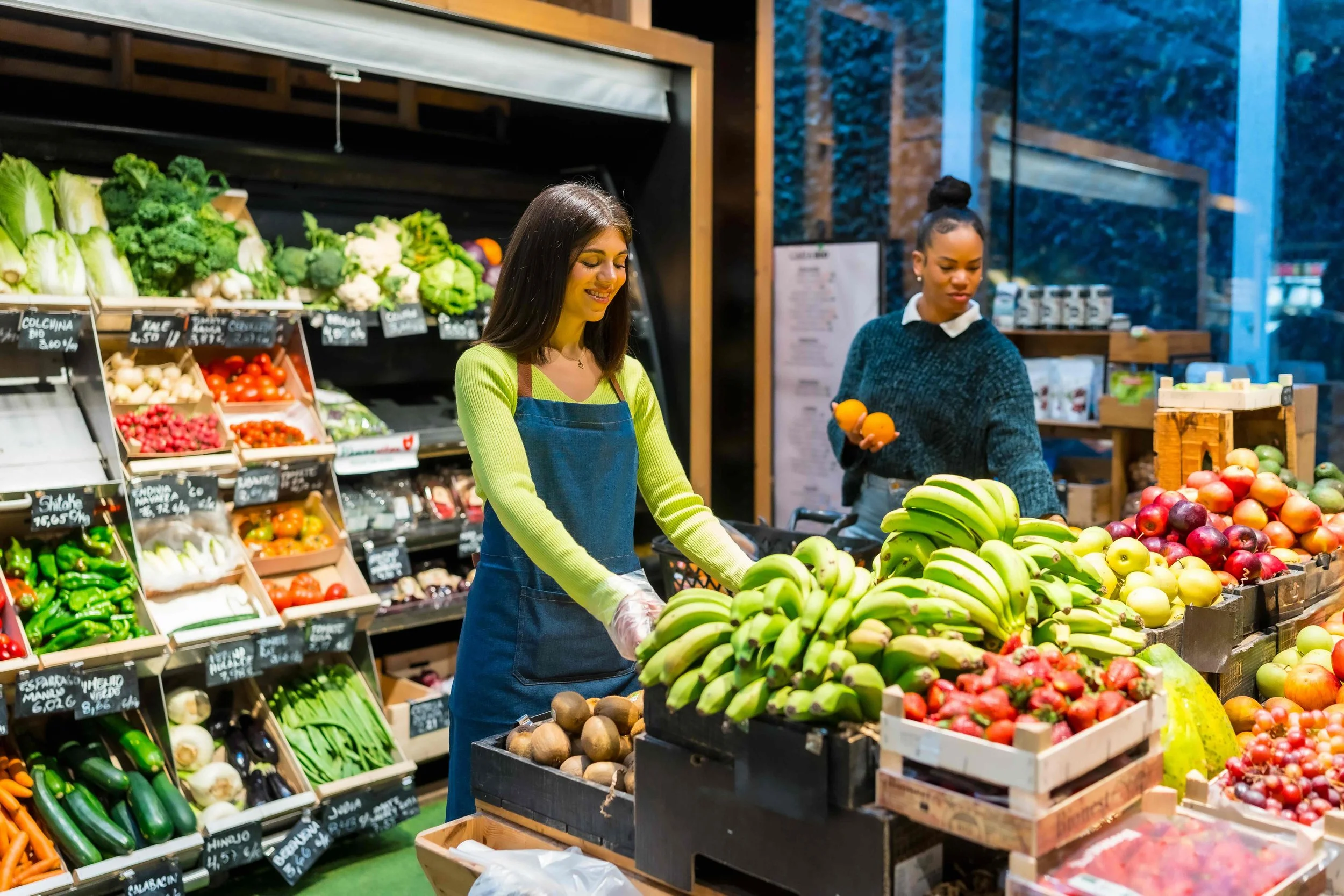 Two women shopping for fruits and vegetables at a grocery store produce section.