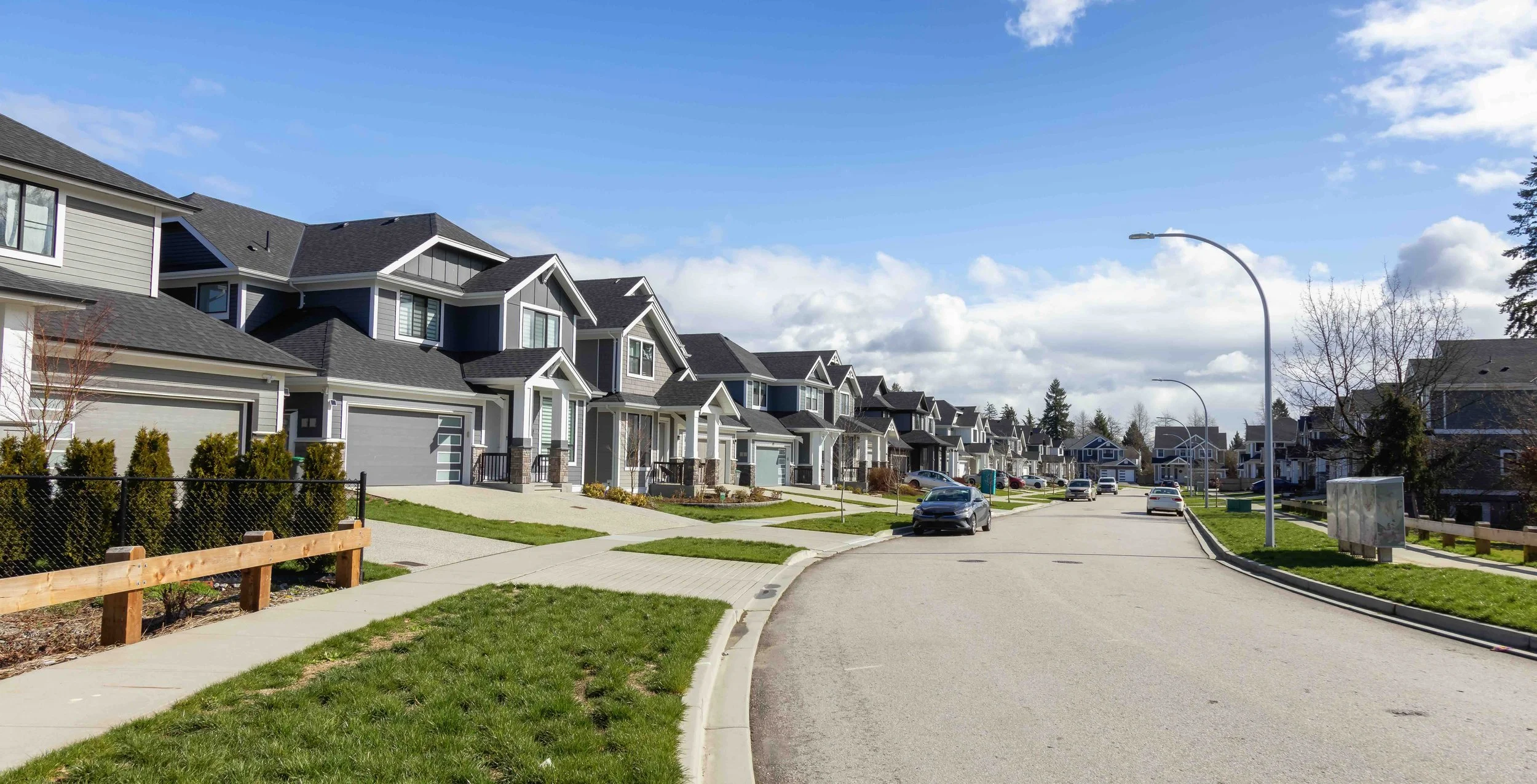 A suburban neighborhood with modern, two-story houses, parked cars, green lawns, and a curved street under a partly cloudy sky.