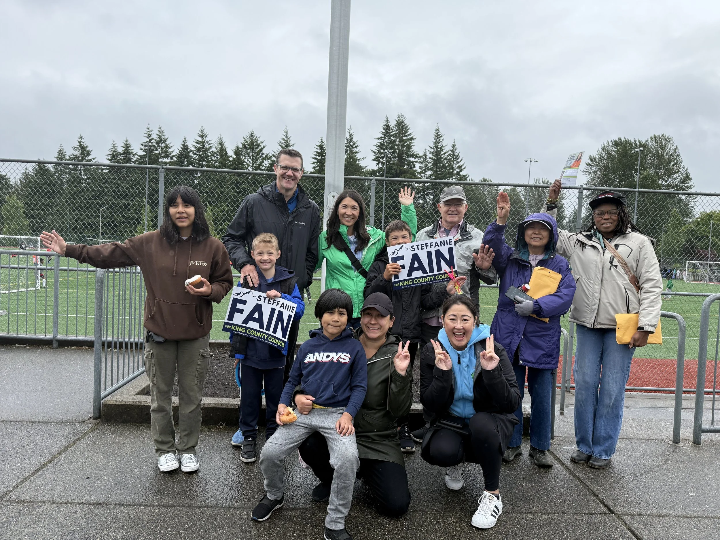 Group of children and adults smiling and posing outdoors at a sports field, some holding campaign signs for Steffanie Fain for King County Council, with overcast weather and wet ground.