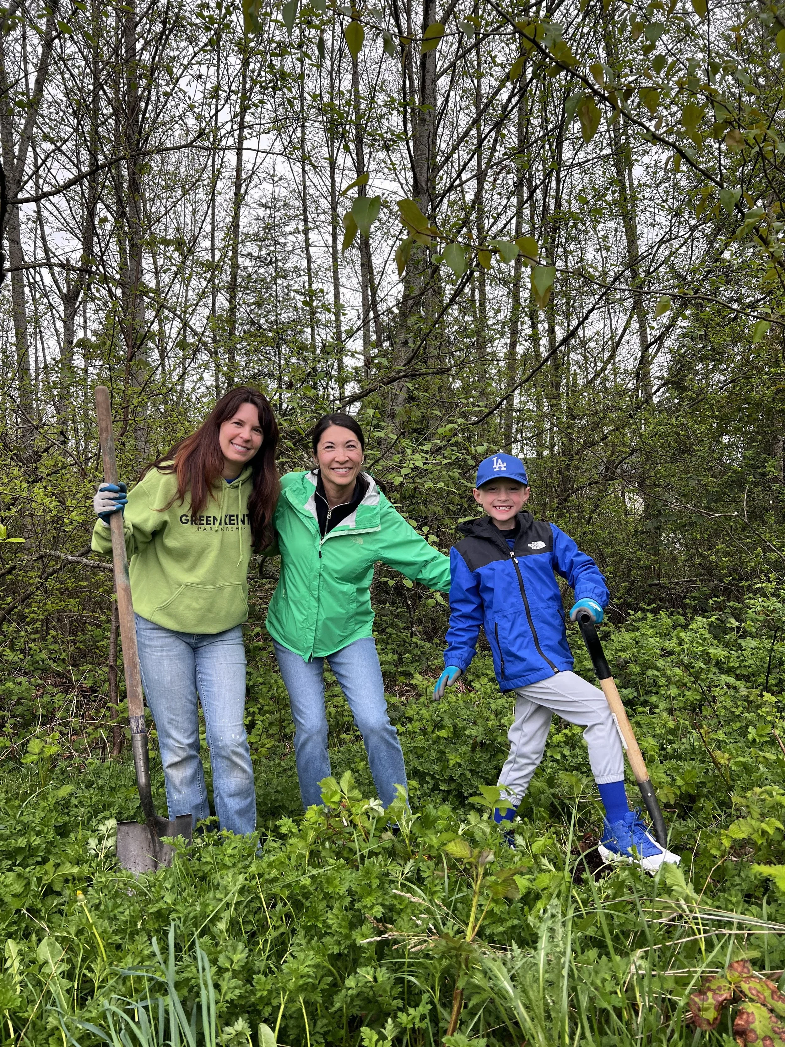 Three people, two women and a boy, standing outdoors in a wooded area, smiling and holding gardening tools amidst green plants and trees.