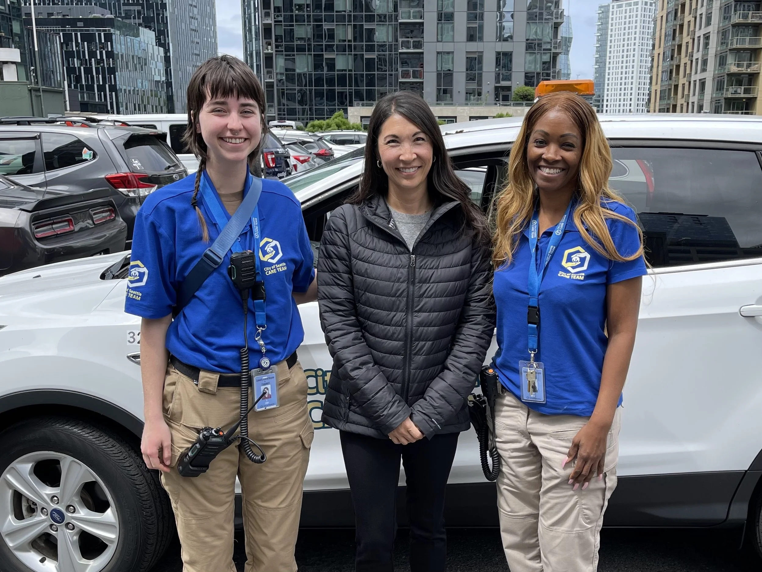 Three women standing in front of a city street police vehicle, smiling at the camera.