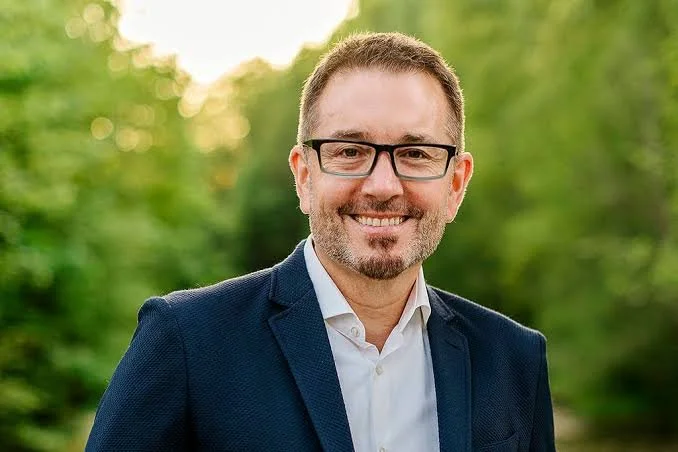 A smiling man wearing glasses and a navy blue suit outdoors with a green, blurred background.
