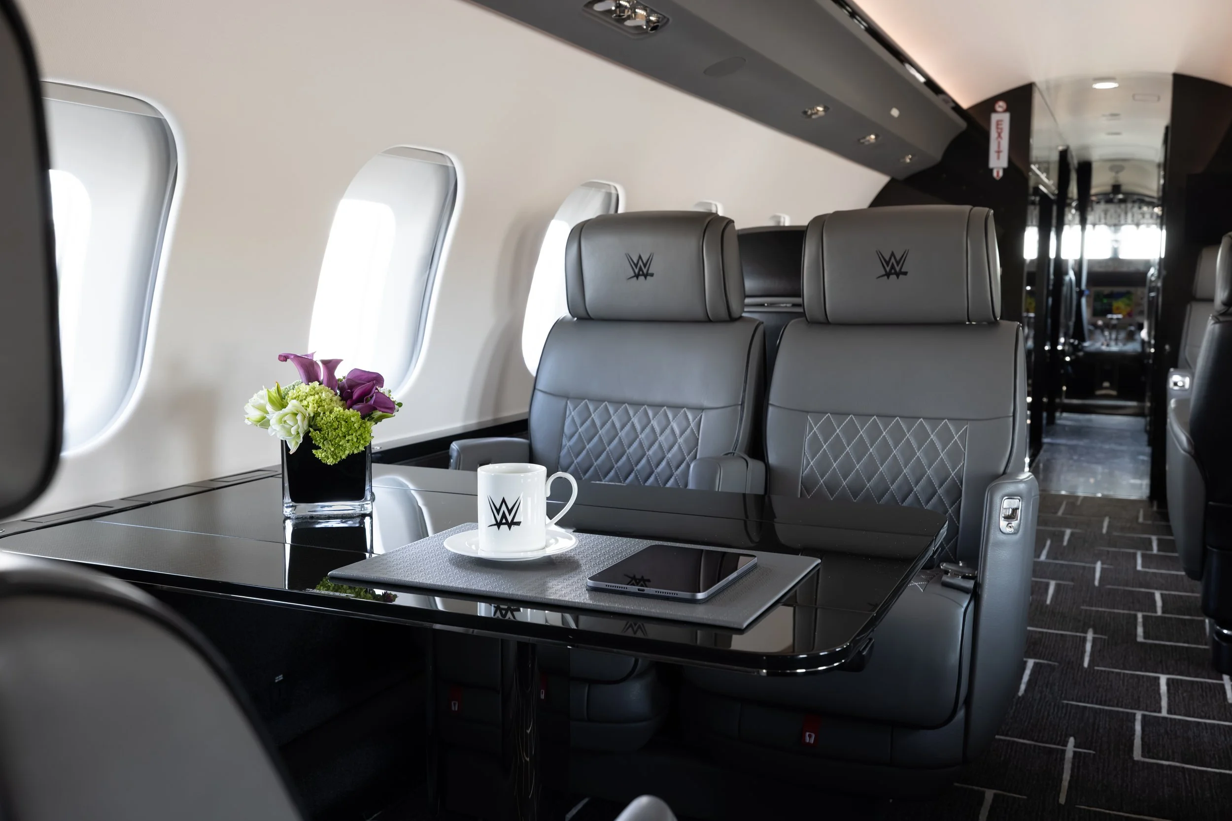 Interior of a Bombardier Global 6500 by Sarah Larranaga Aviation Design with gray leather seats featuring WWE logos, a black table with a vase of flowers, a coffee mug, and a tablet, with windows along the side and modern decor.