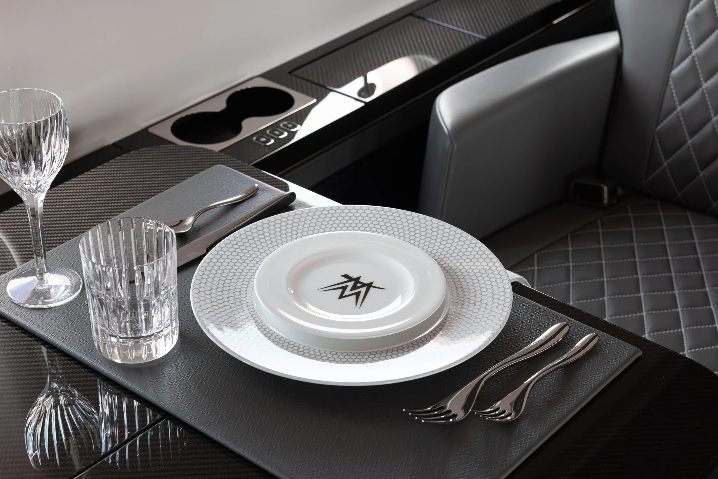 A close-up view of a dining table set with a white plate with black design, a fork and knife, a textured placemat, a crystal wine glass, a water glass, and a gray notebook. In the background, there are black leather chairs and a table with built-in c