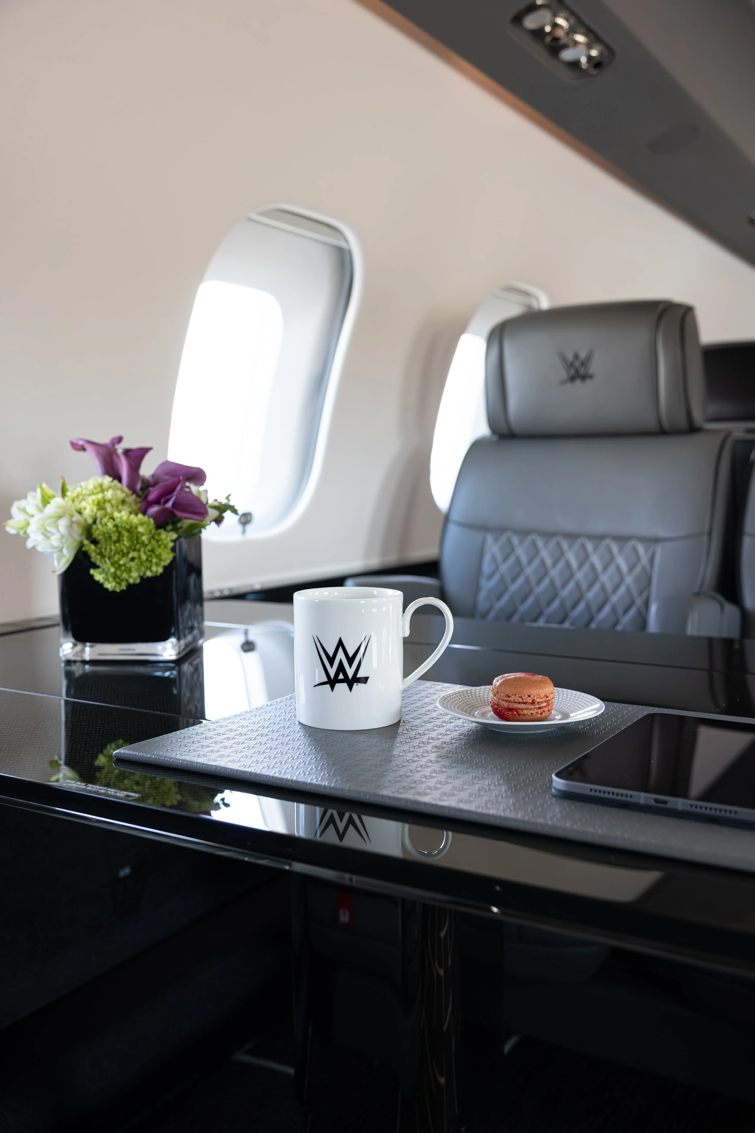 Inside of an airplane cabin with a tray table set with a white coffee mug with the WWE logo, a small plate with a macaroon, a black vase with a bouquet of pink and white flowers, and a smartphone. The background shows a leather seat with the WWE logo