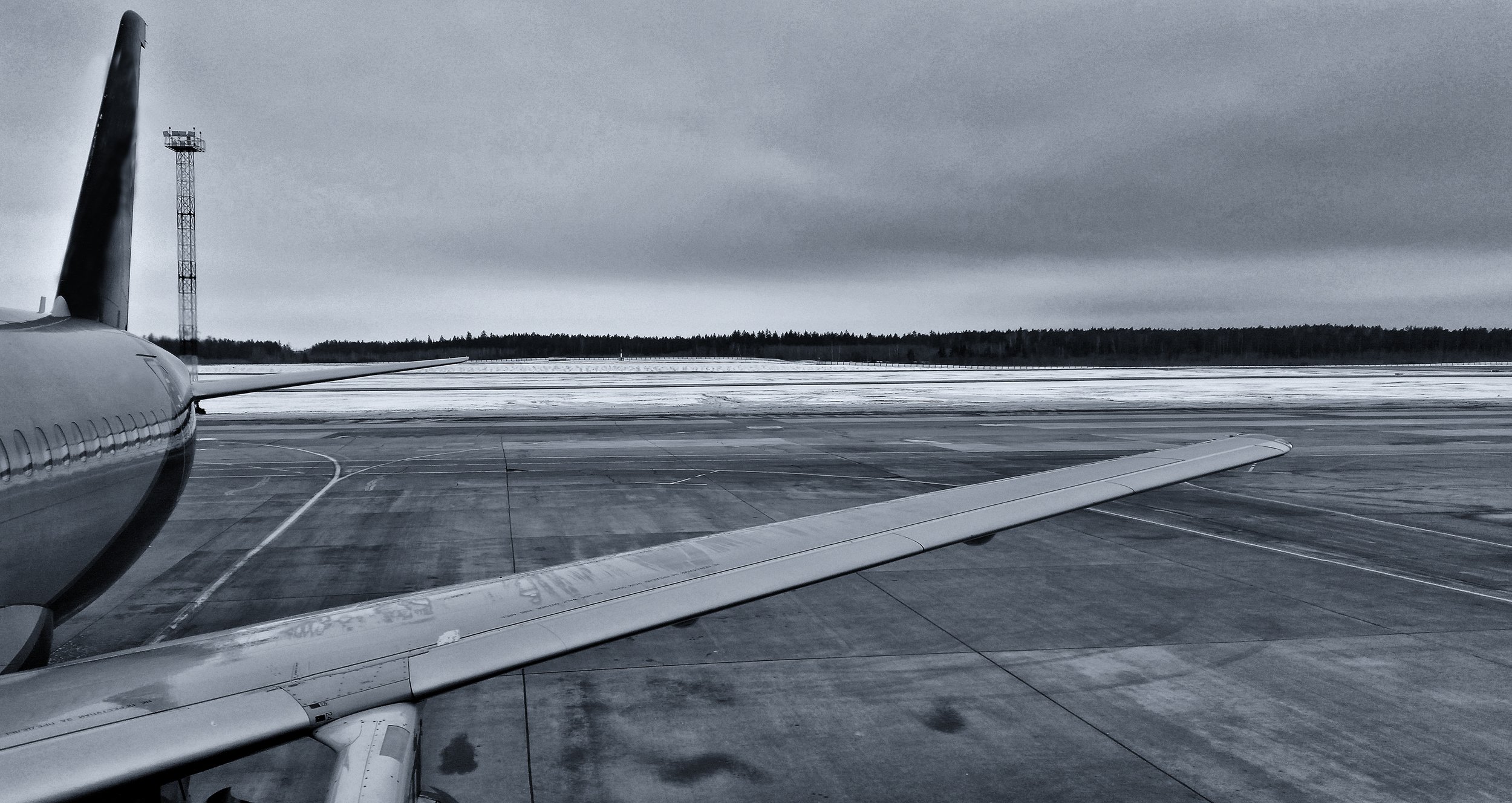 View from an airplane window showing the airplane wing on a snowy airport runway with a cloudy sky and a forest in the background.