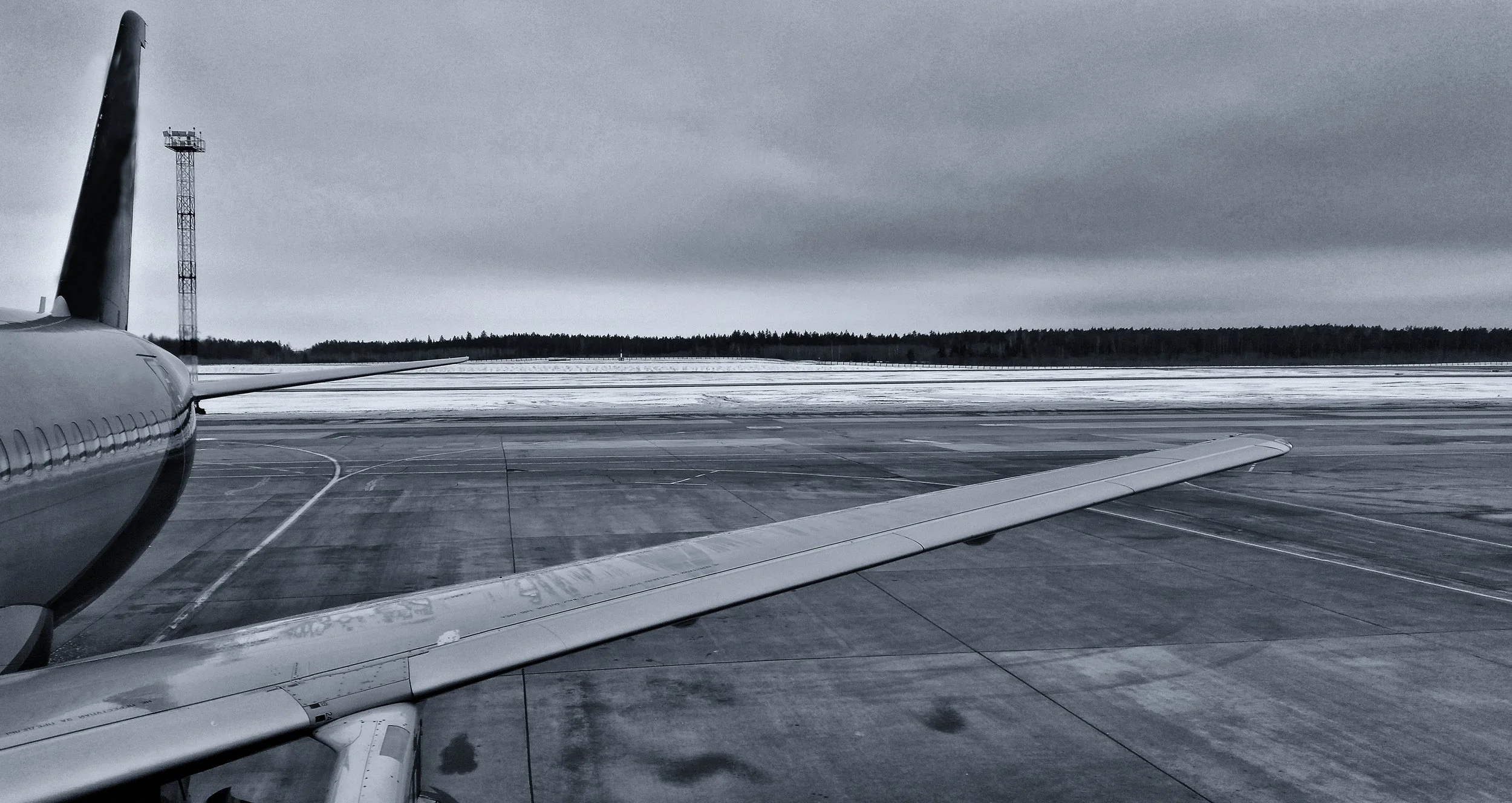 View from an airplane window showing the airplane wing on a snowy airport runway with a cloudy sky and a forest in the background.