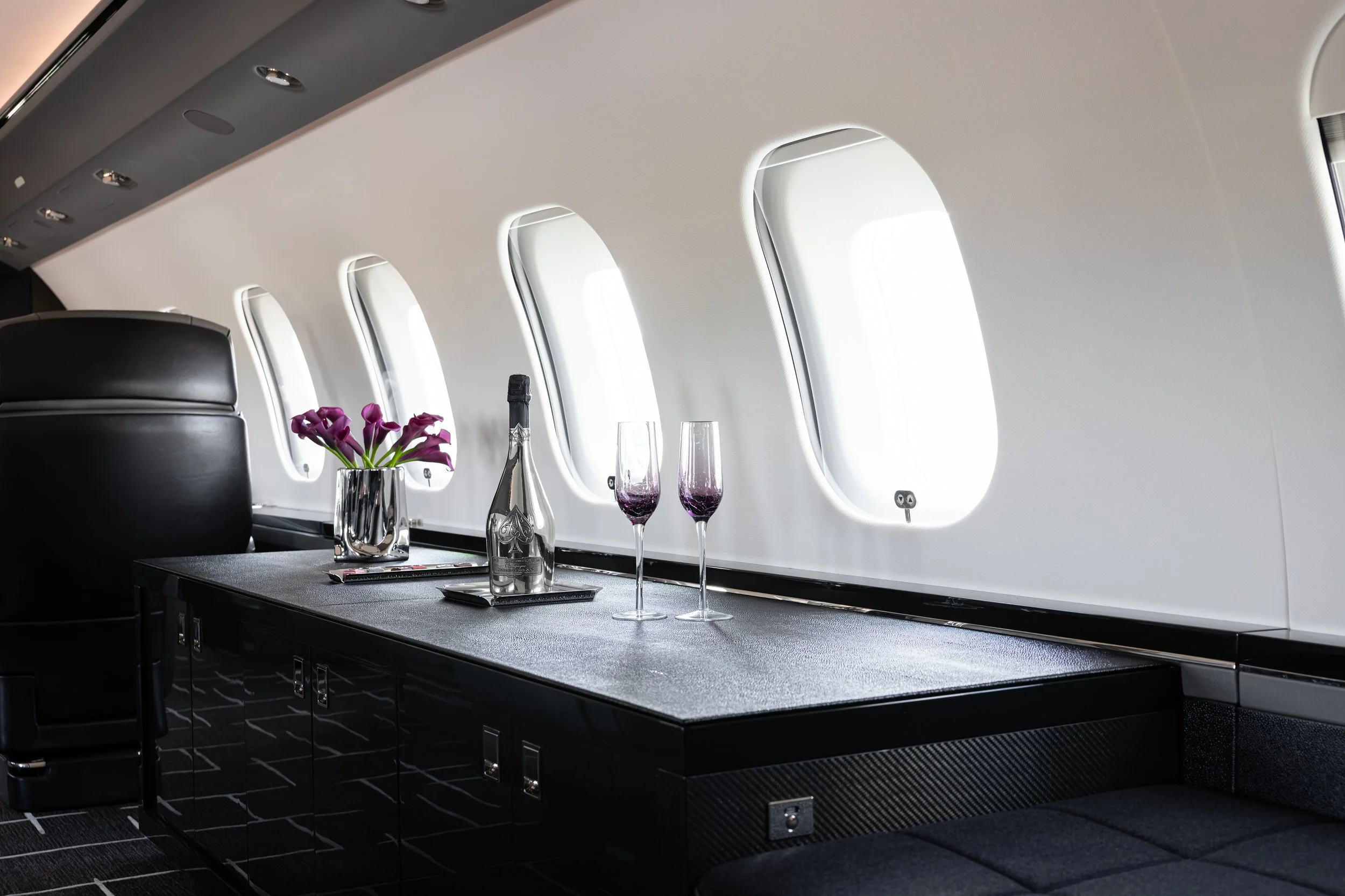 A Bombardier Global 6500 by Sarah Larranaga Aviation Design interior with a table set for a meal. The table has a vase with purple flowers, a bottle of champagne, and two empty champagne flutes. The background shows airplane windows and a black seat.